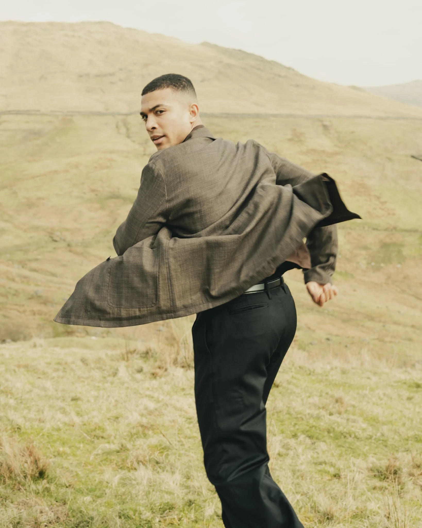 A man in a brown suit and black pants standing in a grassy field, with rolling hills in the background, looking over his shoulder.