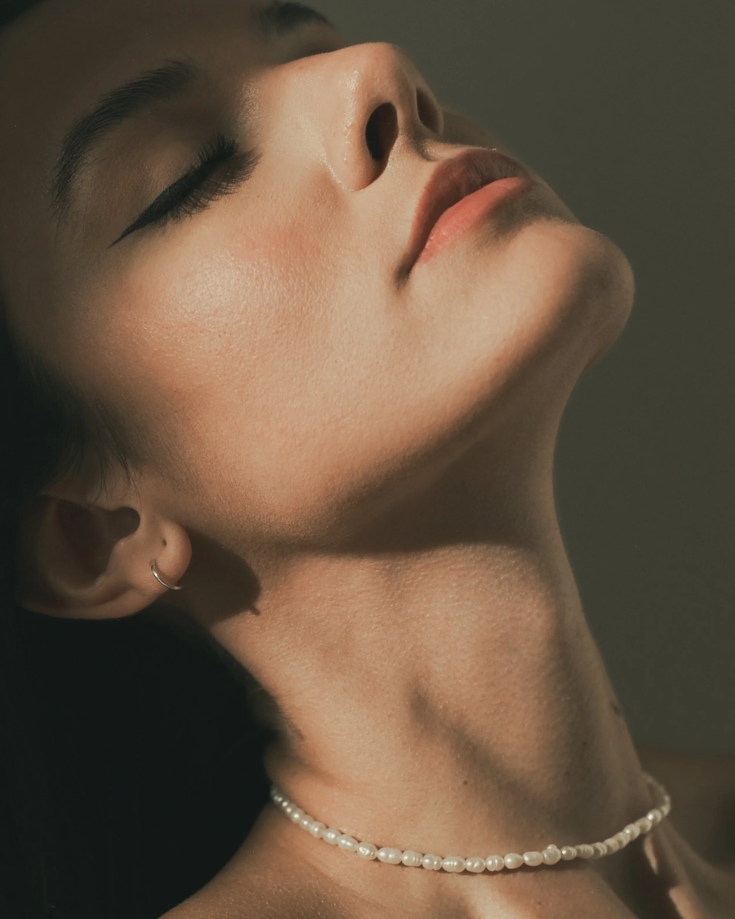 Close-up of a woman's face with closed eyes, wearing a pearl necklace, and having smooth skin and makeup.
