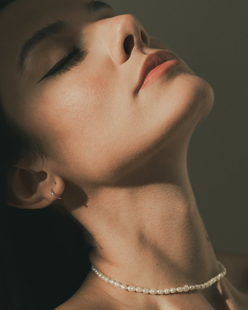 Close-up of a woman's face with closed eyes, wearing pearl jewelry, with smooth skin and styled eyebrows.