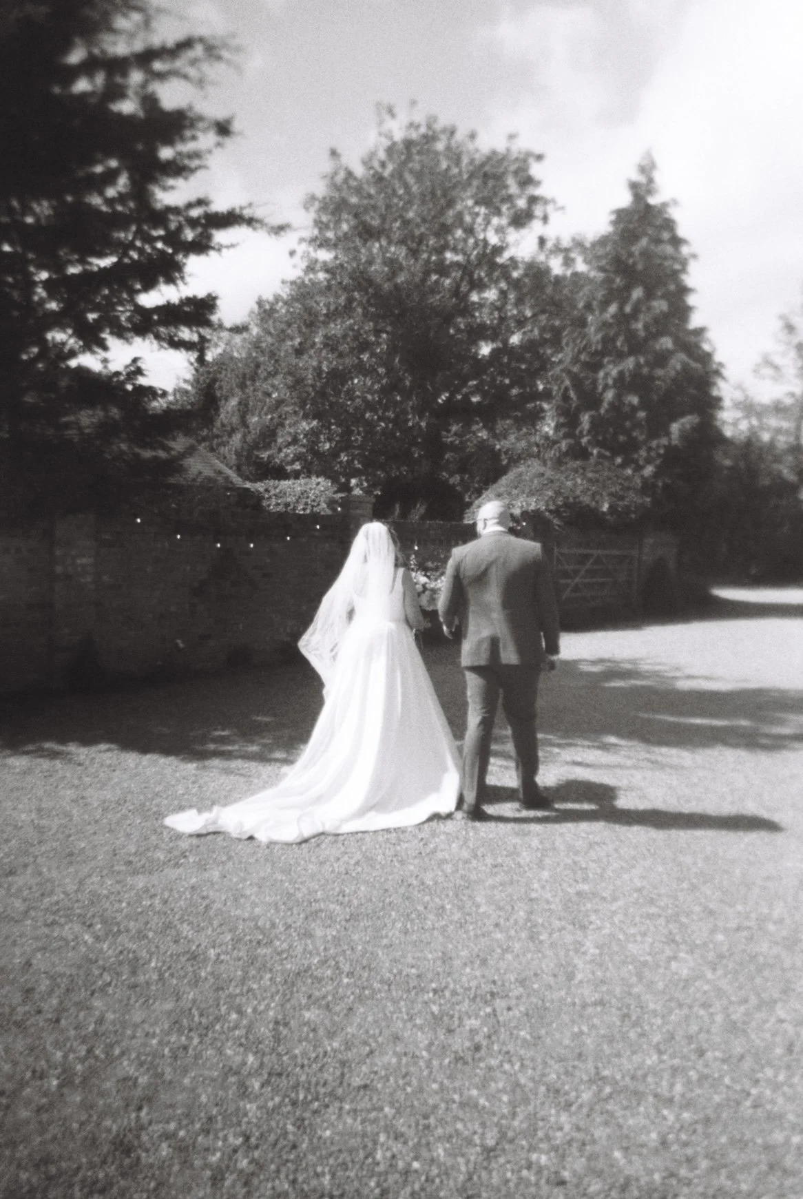 A bride in a white wedding dress and veil and a groom in a suit walking together outdoors on a sunny day, with trees and a brick wall in the background.