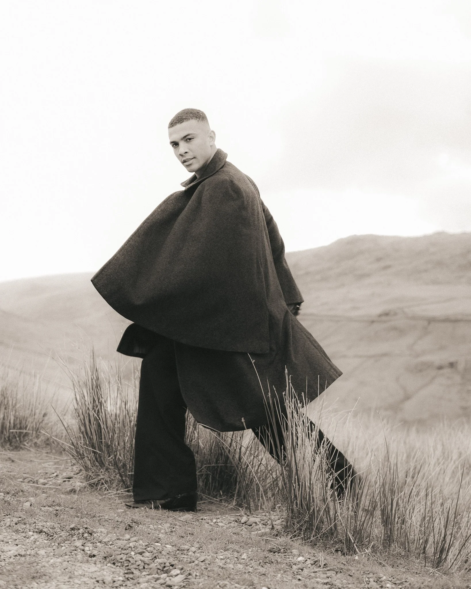 A young man stands outdoors in a dirt area with tall dry grass, wearing a long dark coat, with mountains in the background under an overcast sky.