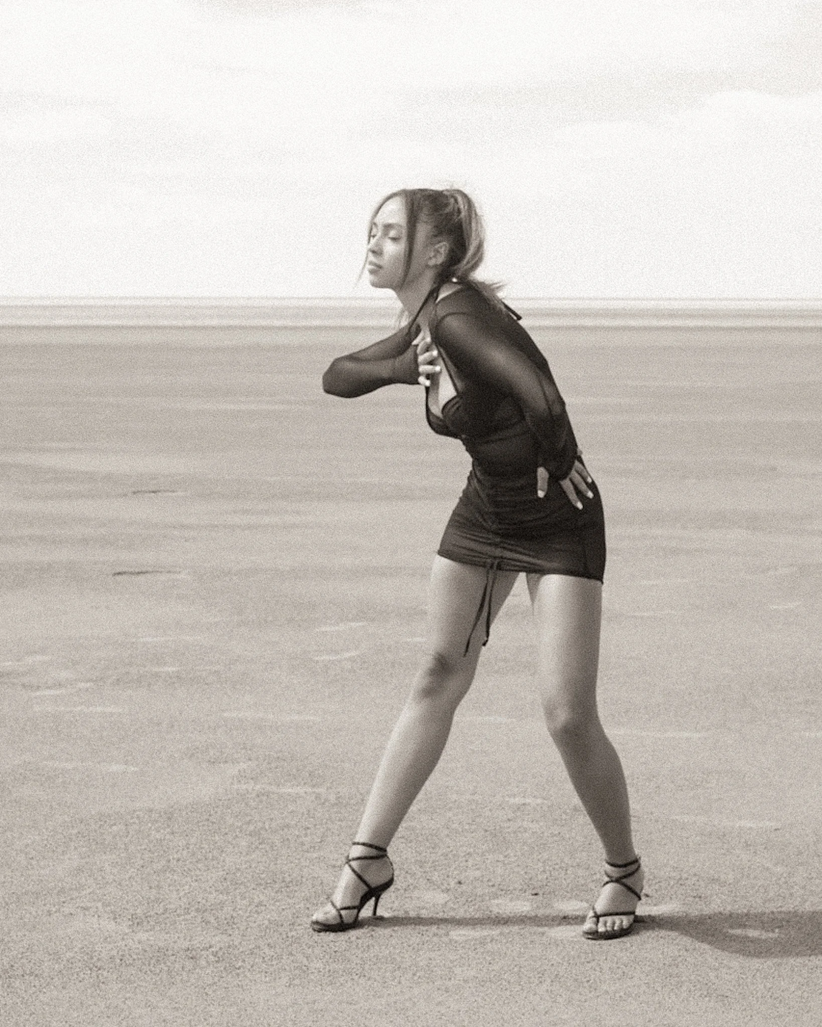 A woman in a black sheer top and black shorts walks on a sandy beach, wearing strappy high heels. Her eyes are closed, and she is holding her chest with one hand while the other hand is placed on her back.