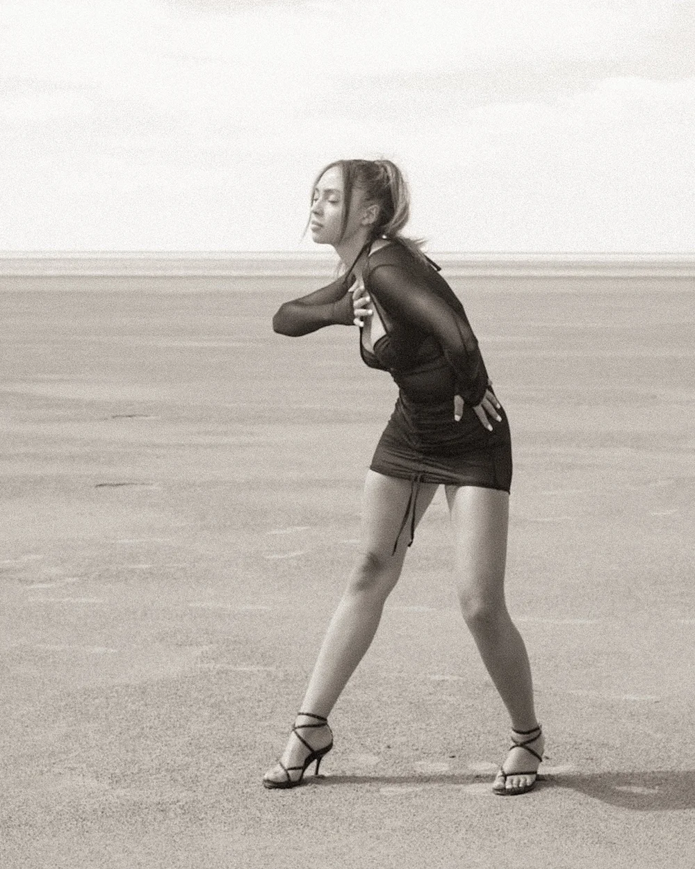 A woman in a black sheer top and black shorts walks on a sandy beach, wearing strappy high heels. Her eyes are closed, and she is holding her chest with one hand while the other hand is placed on her back.