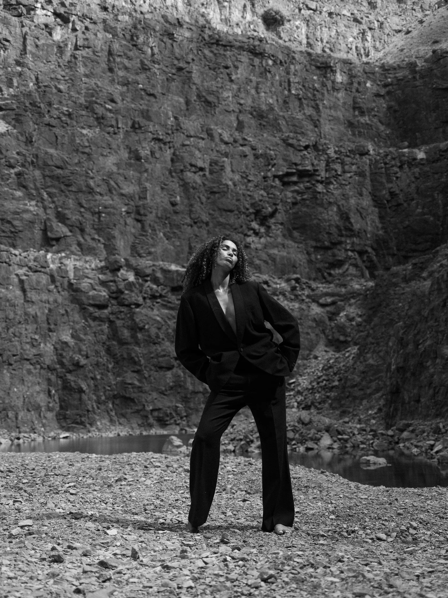A woman with curly hair wearing a dark blazer and pants stands with her hands in her pockets near a rocky riverbank with a canyon in the background.