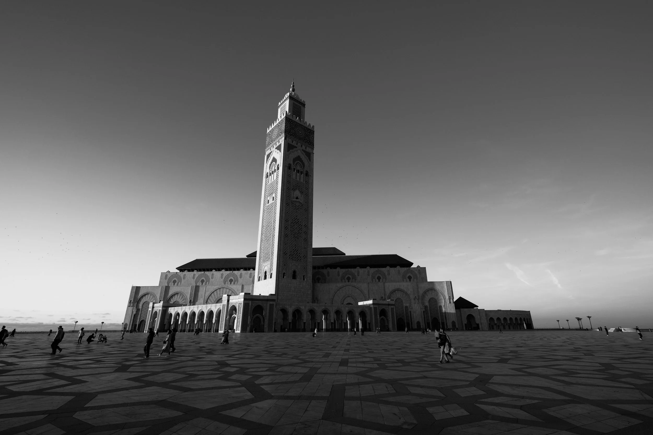A tall, ornate tower with a square base and decorative arches, located in a large open plaza with patterned tile flooring. People are walking and sitting in the plaza, and the sky is clear.