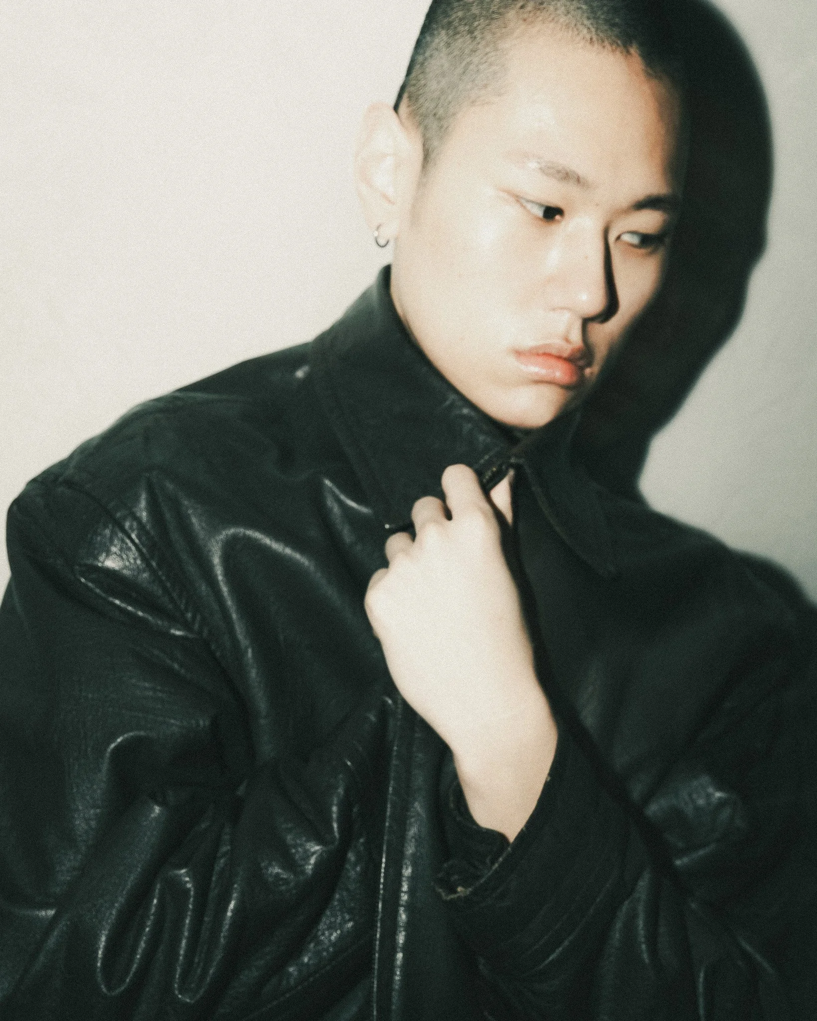 Close-up of a woman with a buzz cut hairstyle, wearing a black leather jacket and hoop earrings, looking down and to the side with a neutral expression.