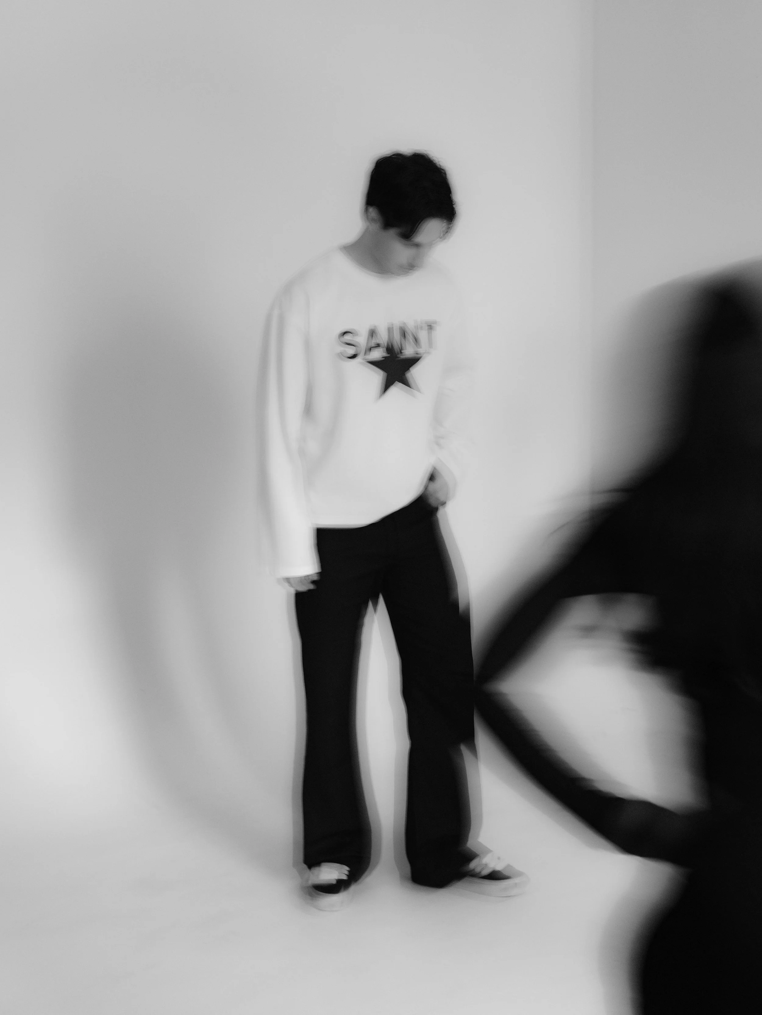 A person with dark hair stands against a plain white wall, wearing a long-sleeve white shirt with the words 'SAINT' and a star printed on it, along with loose black pants and sneakers.