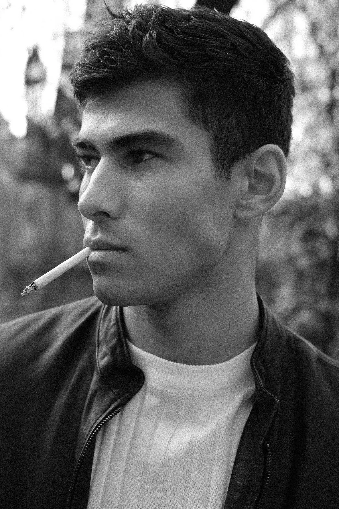 Black and white close-up photo of a young man with dark hair, wearing a leather jacket and white shirt, holding a cigarette in his mouth, looking to the side outdoors.