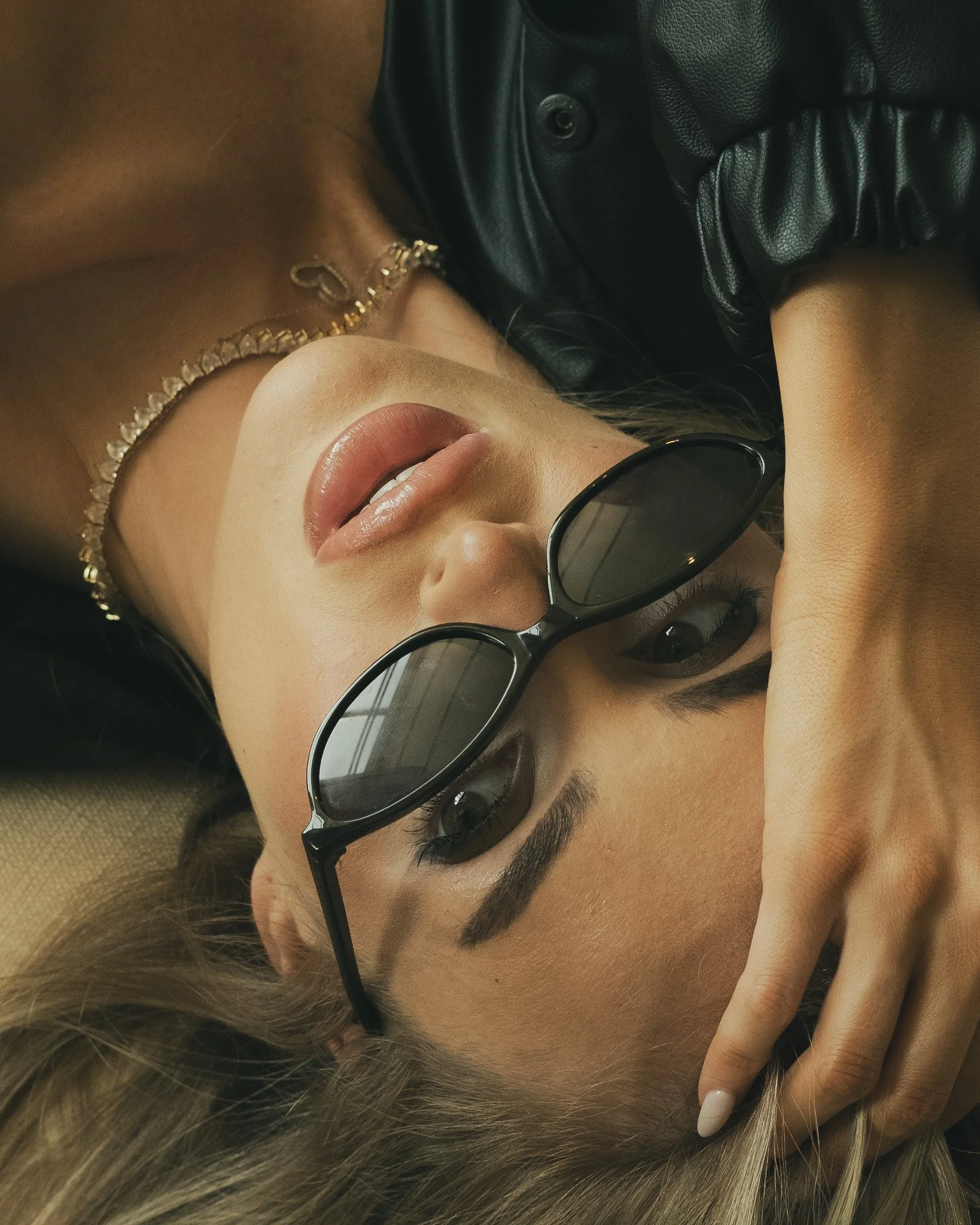 Close-up of a woman lying down with blonde hair, wearing round black sunglasses, a gold necklace, and a black leather jacket.