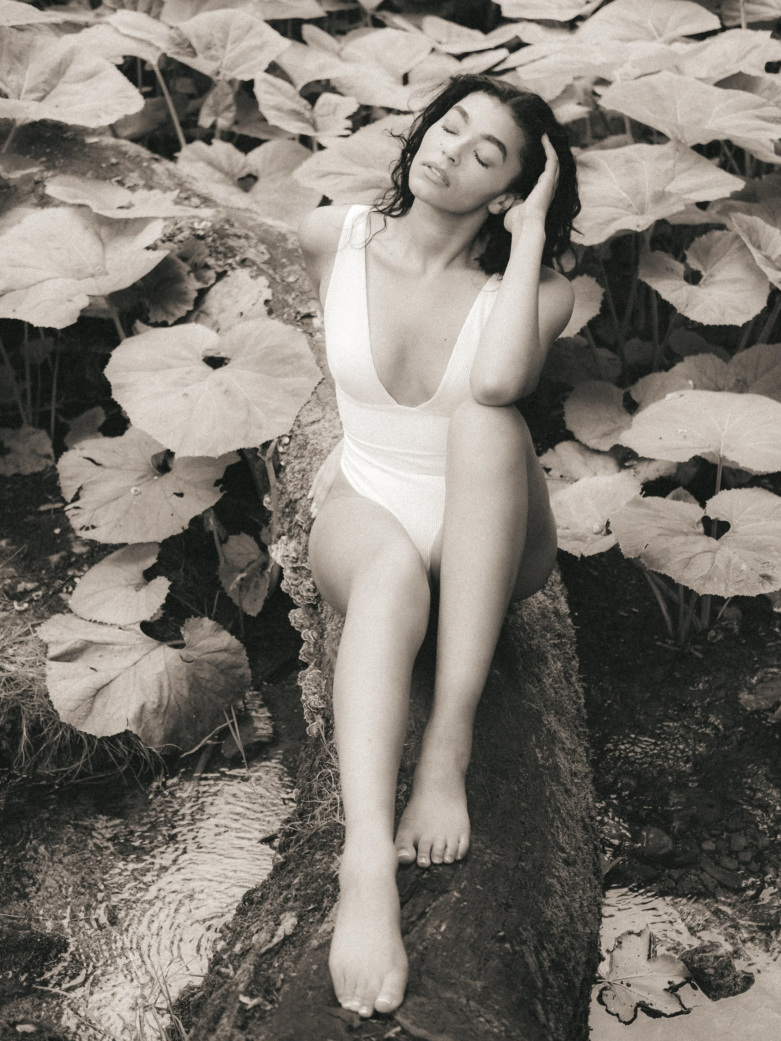 Woman with closed eyes, sitting on a fallen tree trunk among large leaves, with one hand on her head and the other resting on her knee, wearing a white sleeveless top.