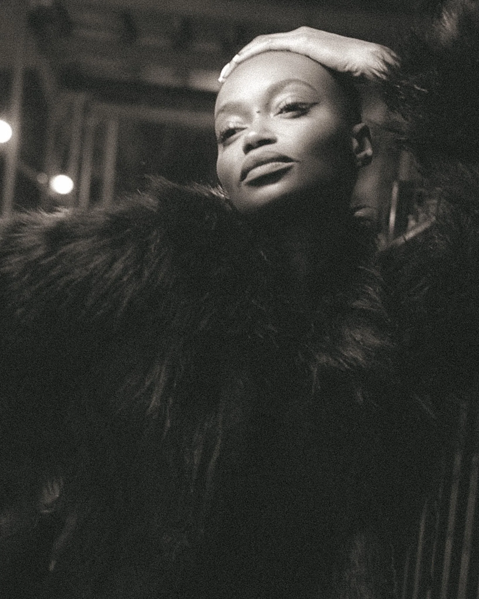 Black and white photograph of a woman with closed eyes, wearing a headscarf and a fur coat, with a blurred background of lights.
