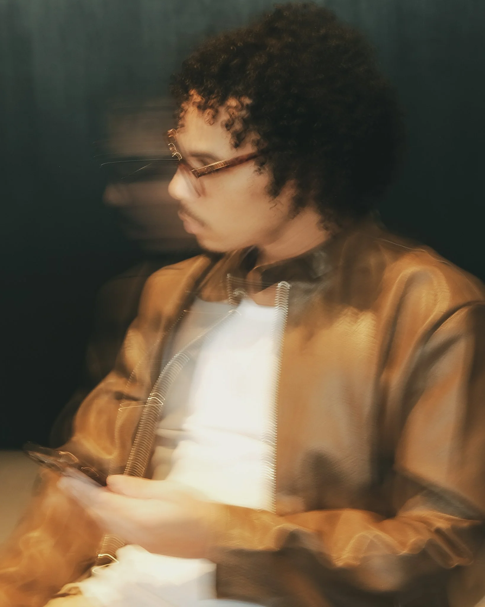 A person with curly hair and glasses, wearing a brown jacket, looking down with a blurred background.