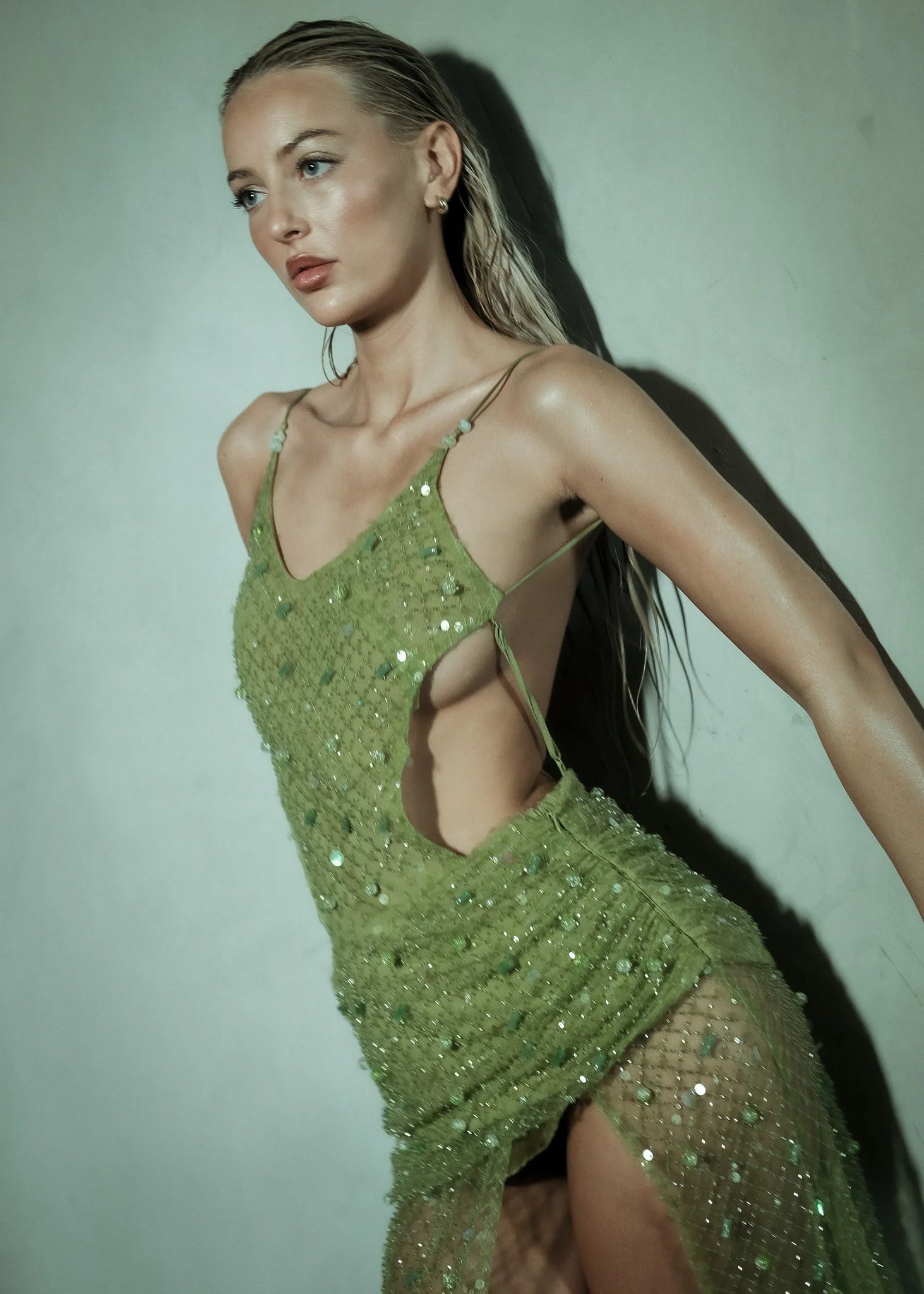 A woman with wet blonde hair, wearing a revealing, green, sequined dress with cutouts, standing against a plain wall.