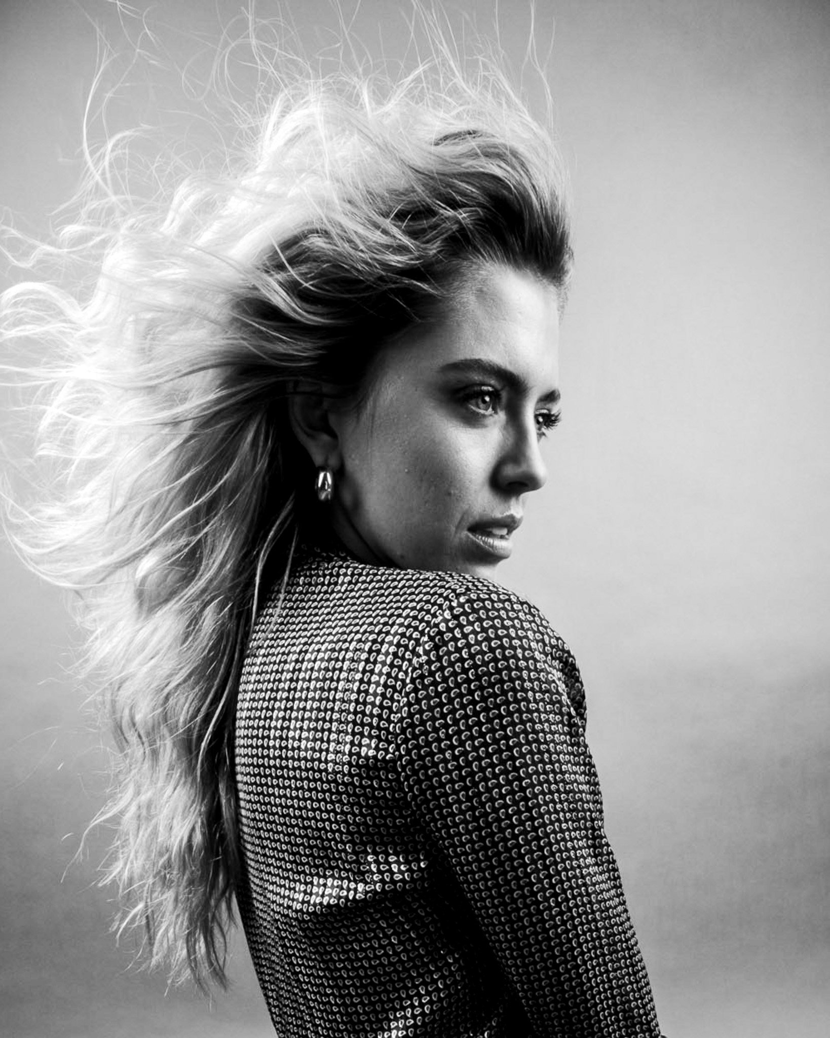 Black and white close-up of a woman with long, wavy hair flowing to the left, wearing a patterned top, with a focus on her face and hair, against a plain background.