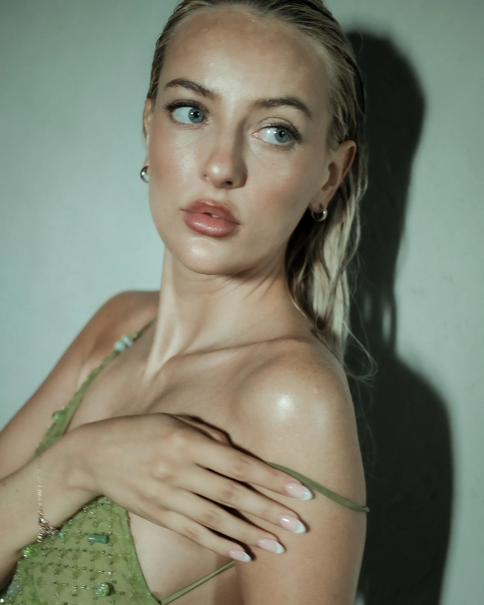 A woman with blonde hair brushed back, wearing a green dress with beaded embellishments, earrings, a necklace, and a bracelet. She is touching her shoulder with her hand and looking slightly off to the side.