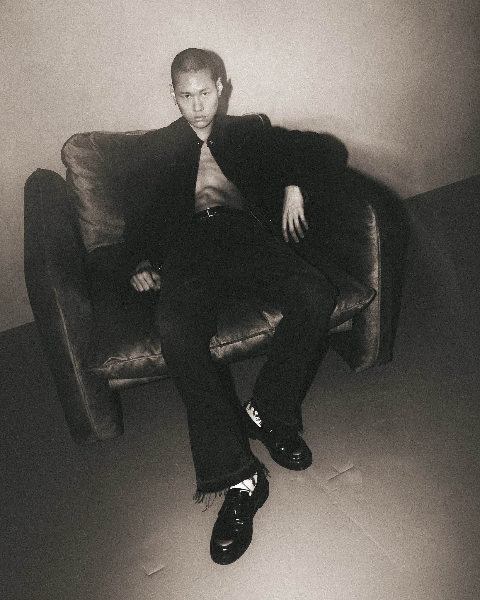Person sitting on couch in black jacket and pants, wearing black shoes and white socks, with crossed legs, in a minimalist room with plain wall and tiled floor, captured in black and white.