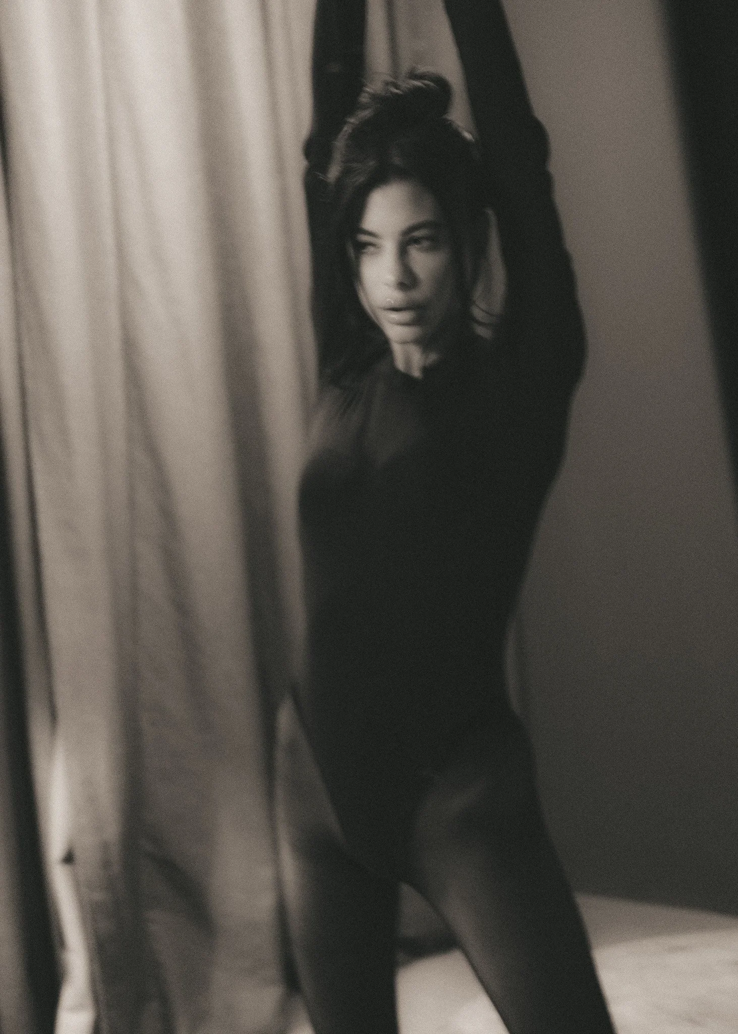 A woman with dark hair styled in curls looking into a mirror, with her hand behind her head, in a black-and-white photo.