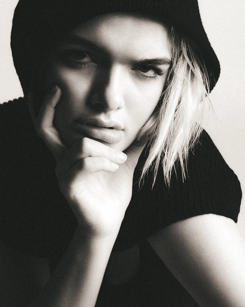 Black and white portrait of a woman with platinum blonde hair, wearing a dark hoodie with the hood up, looking at the camera with her hand touching her face.