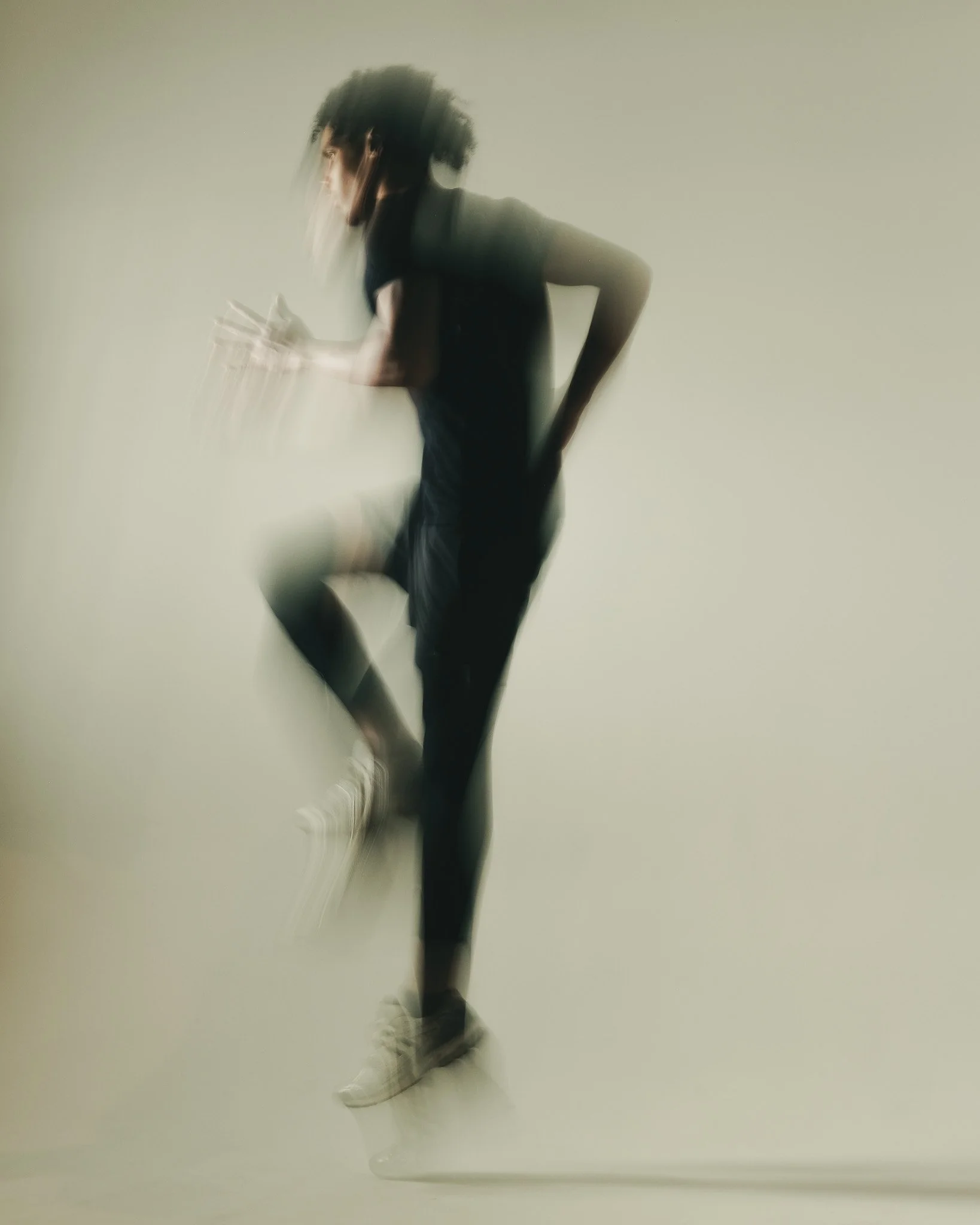 A person with curly hair running or jumping, wearing dark clothing and white sneakers, against a plain light-colored background.