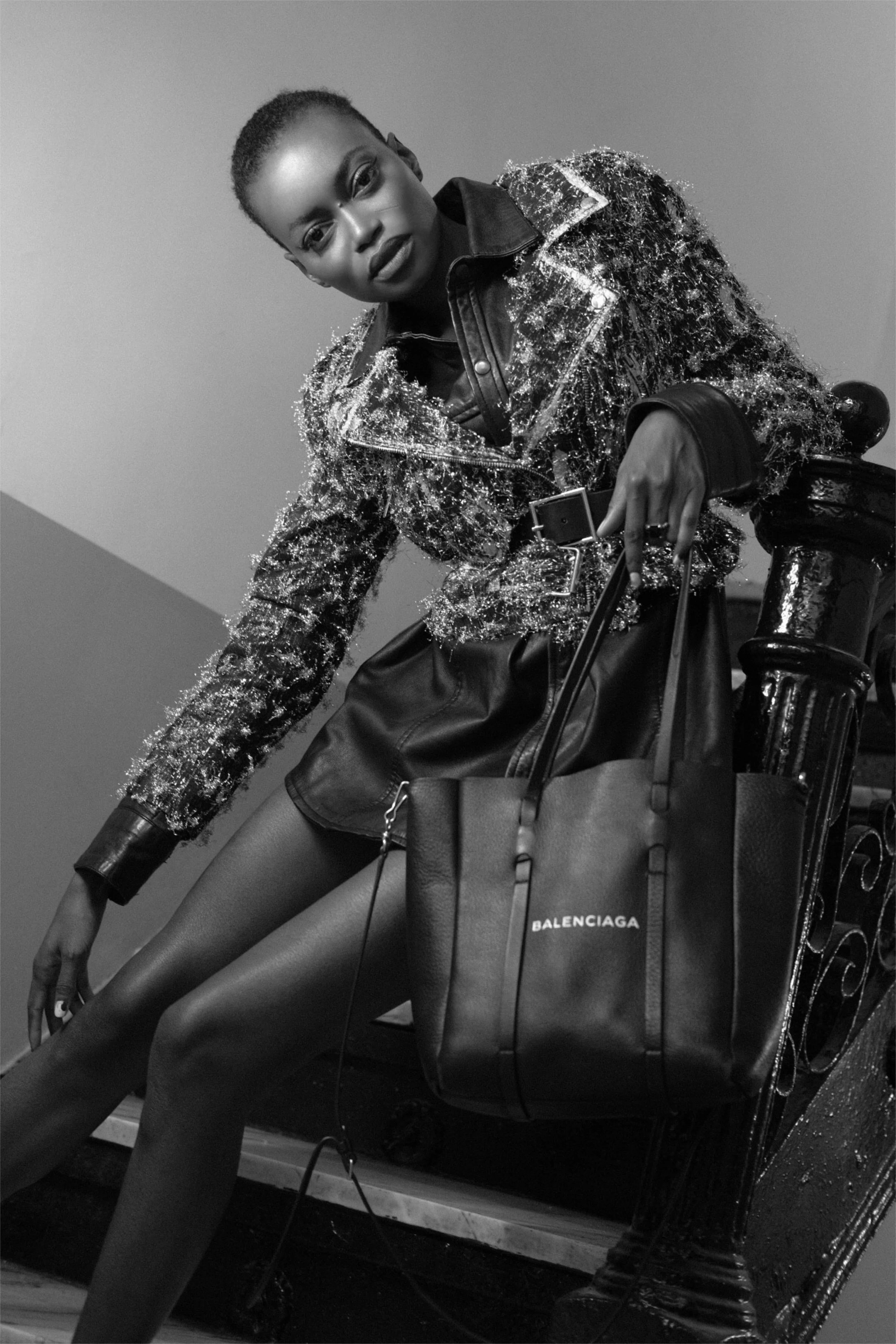 A woman with short hair wearing a sparkling jacket, leather skirt, and tights sitting on a bicycle, holding a Balenciaga shopping bag.