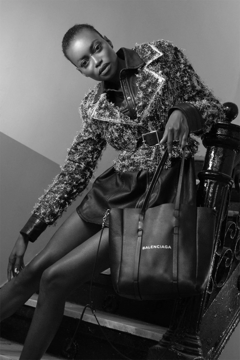 A woman with short hair wearing a sparkling jacket, leather skirt, and tights sitting on a bicycle, holding a Balenciaga shopping bag.