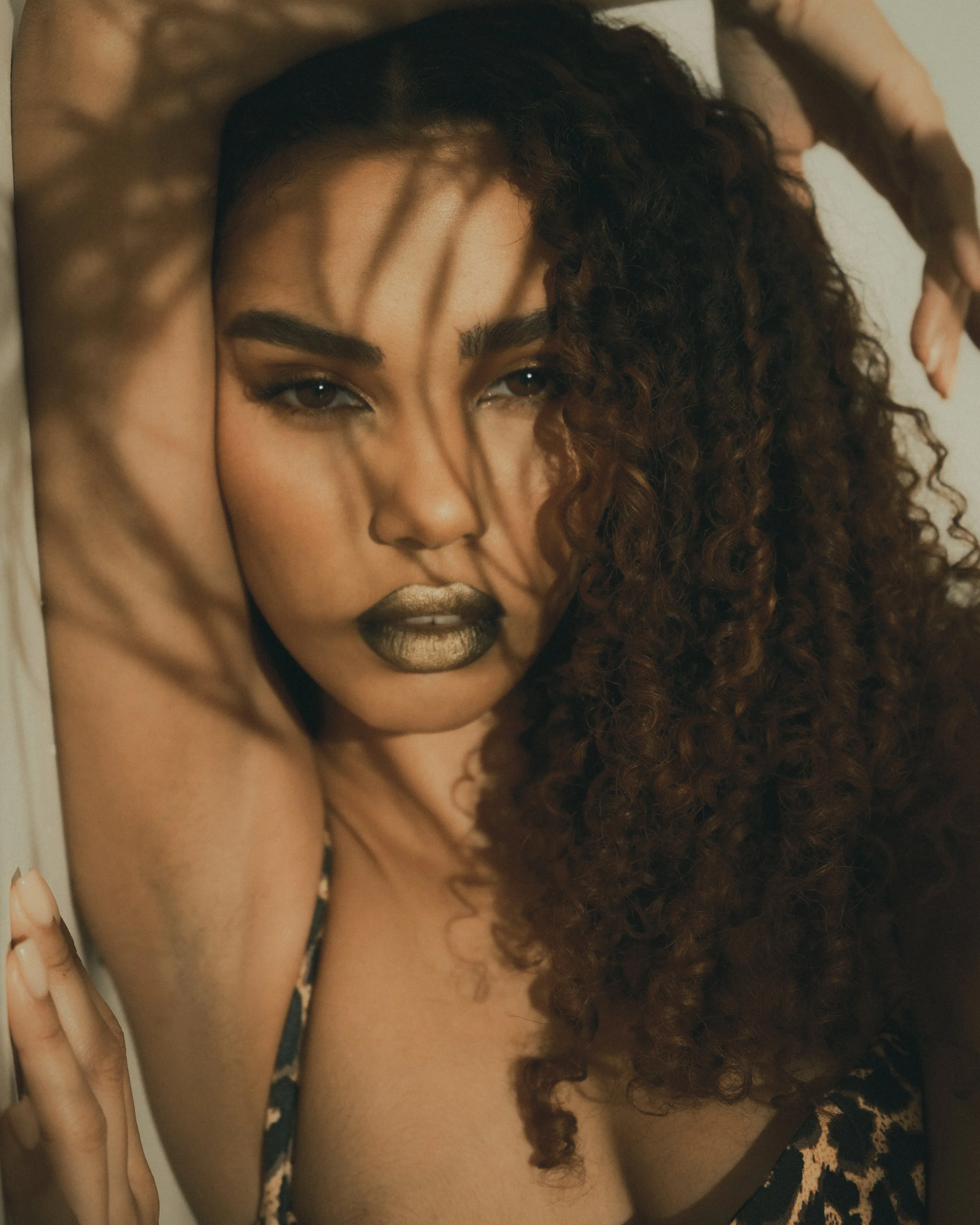 Close-up of a woman with dark curly hair, gazing at the camera, with her hand resting on her head, and golden lipstick.