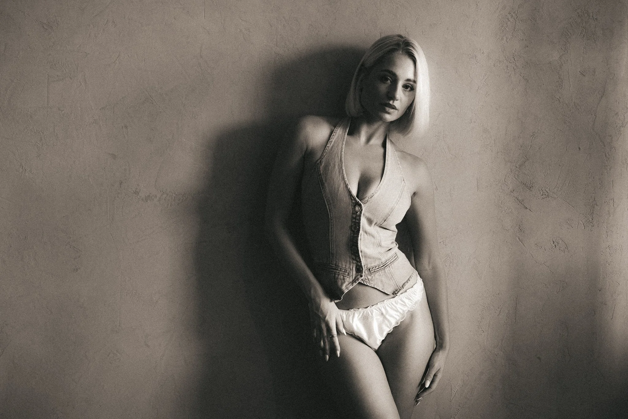 A black-and-white photo of a woman with short blond hair leaning against a textured wall, wearing a sleeveless denim vest and shorts.