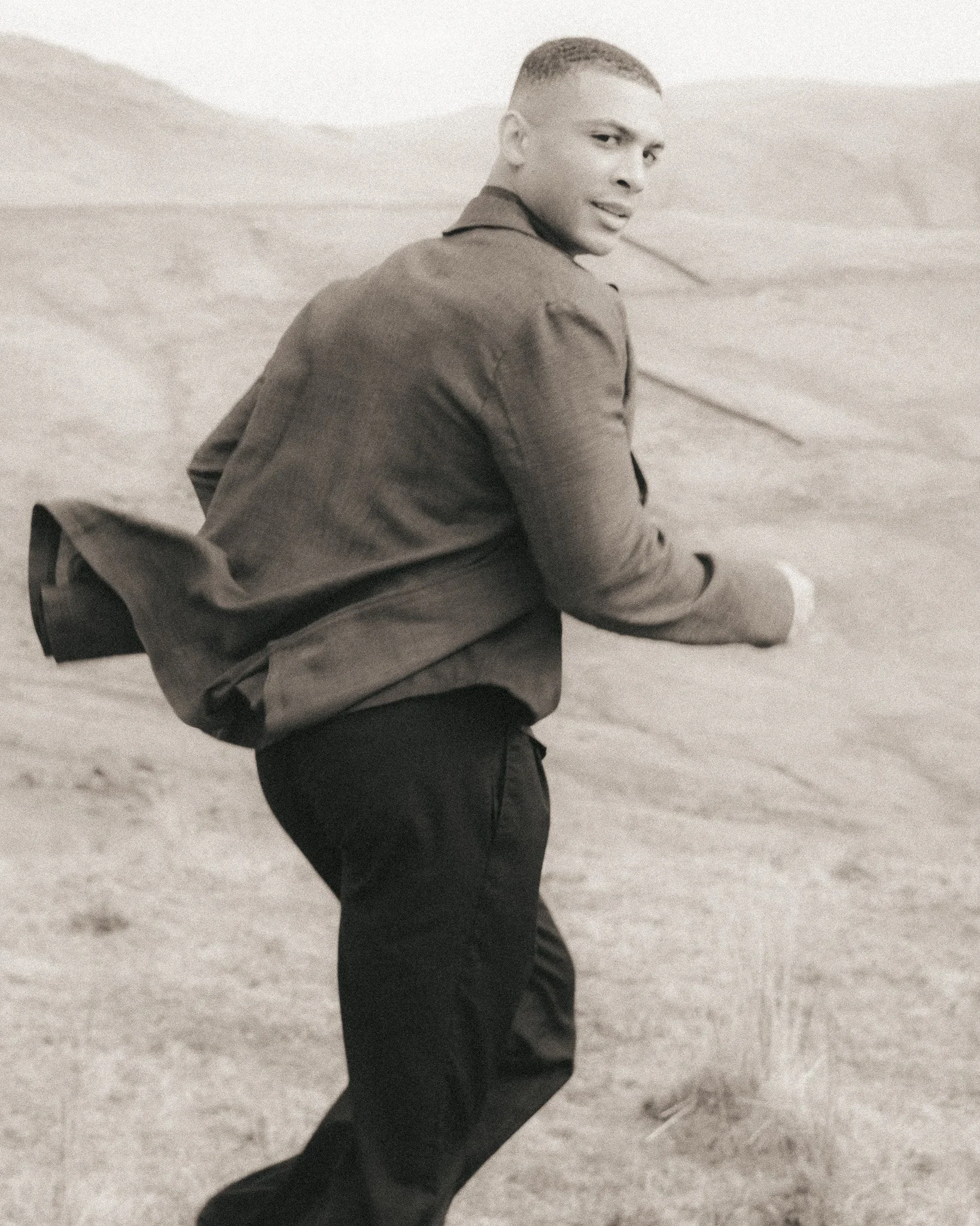 A young man in a jacket and dark pants sitting on a chair outdoors on a sandy surface, turned slightly to the side and looking at the camera.