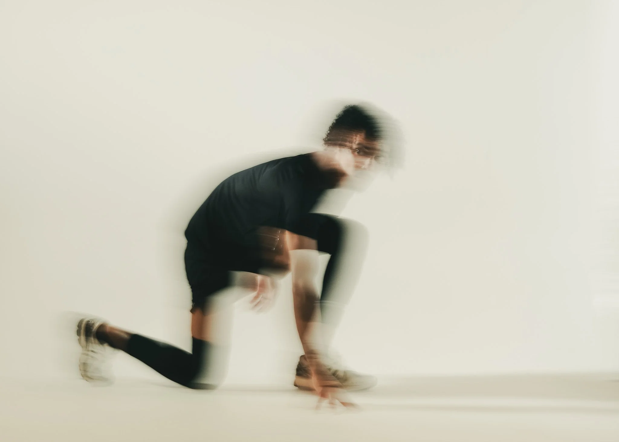 A person is captured mid-motion, performing a handstand or hand-support movement against a plain light-colored wall. The person is wearing dark clothing and sneakers, with their head and upper body slightly blurred due to movement.