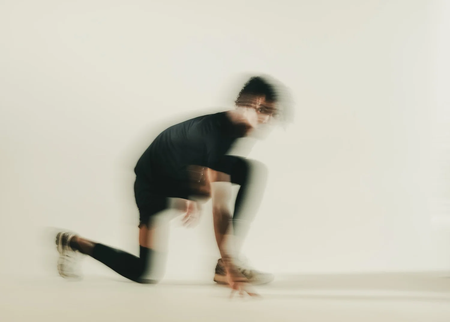 A person with dark curly hair in a black shirt and black pants practicing handstands against a white wall, with their legs bent and one hand on the ground.