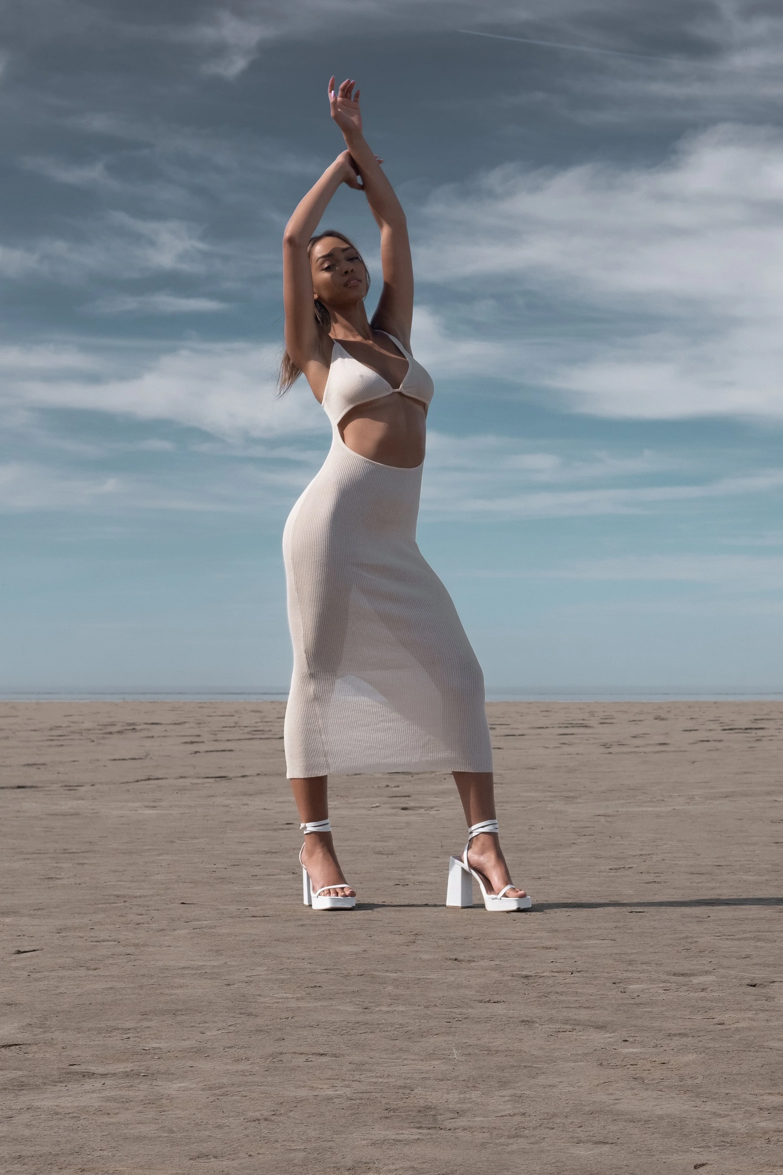 A woman in a beige dress and high heels standing on a wide, flat desert area under a cloudy sky, with her arms raised above her head in a graceful pose.