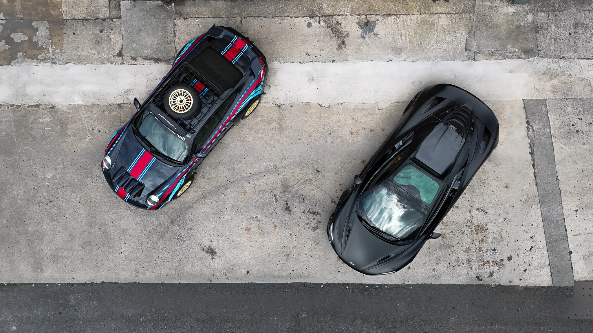 An aerial view of two parked cars on a concrete parking lot, one with racing stripes and a spare tire on top and the other black.