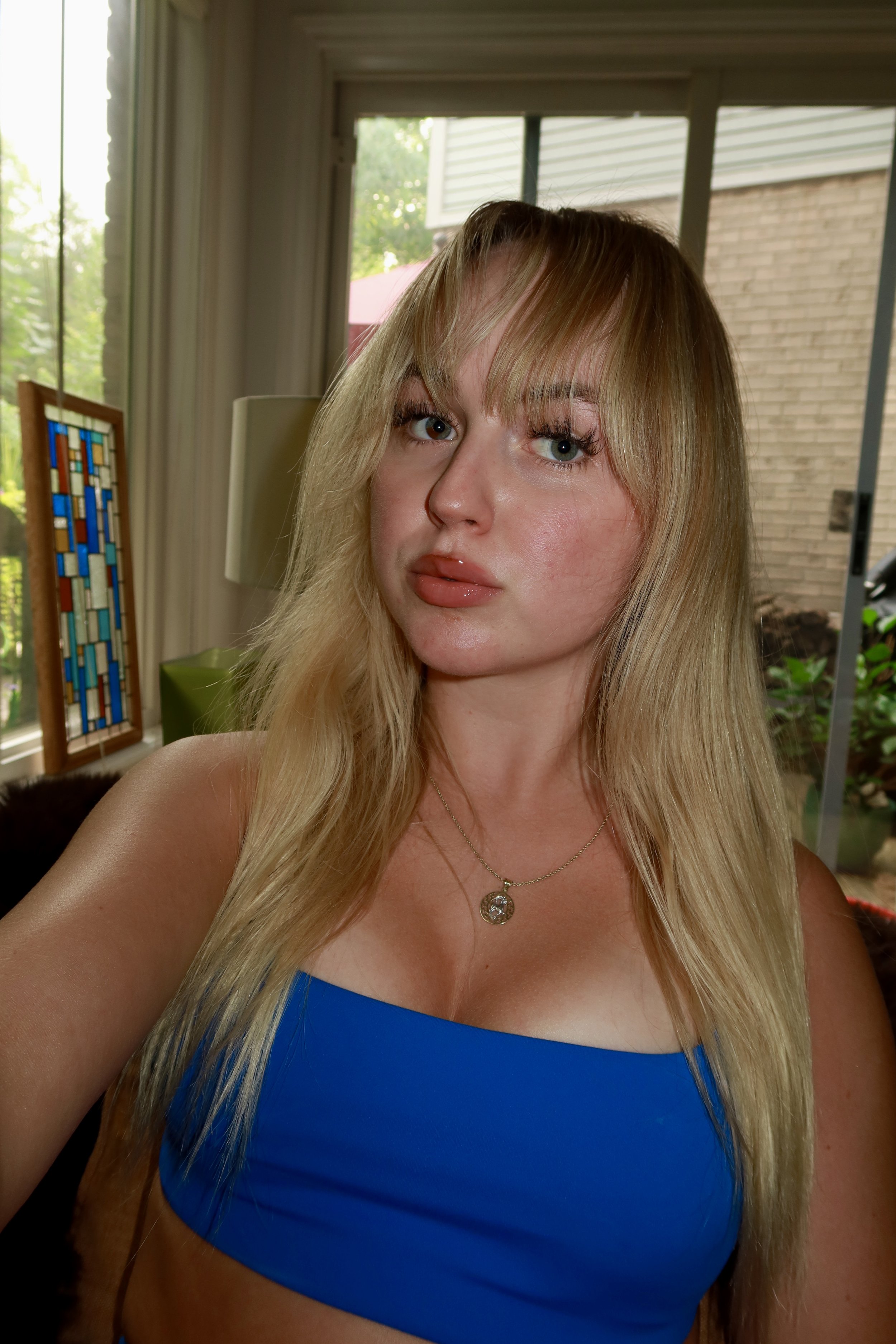 A woman with long blonde hair, blue eyes, and wearing a blue strapless top and a necklace, taking a selfie indoors with a living room and backyard visible through windows behind her.