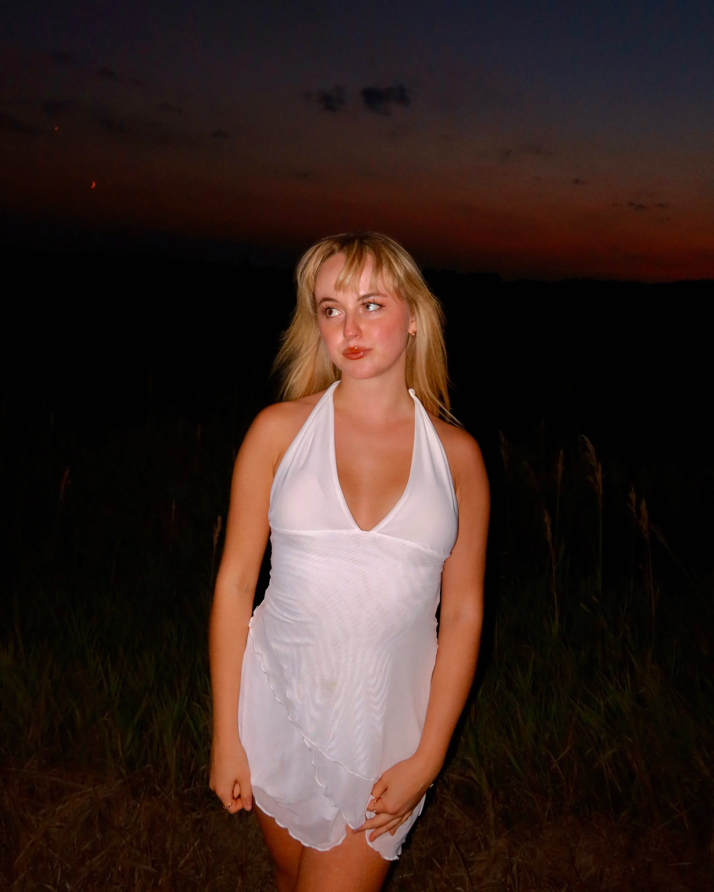 A woman with blonde hair standing outdoors at dusk, wearing a white dress, with a dark sky and faintly visible crescent moon in the background.