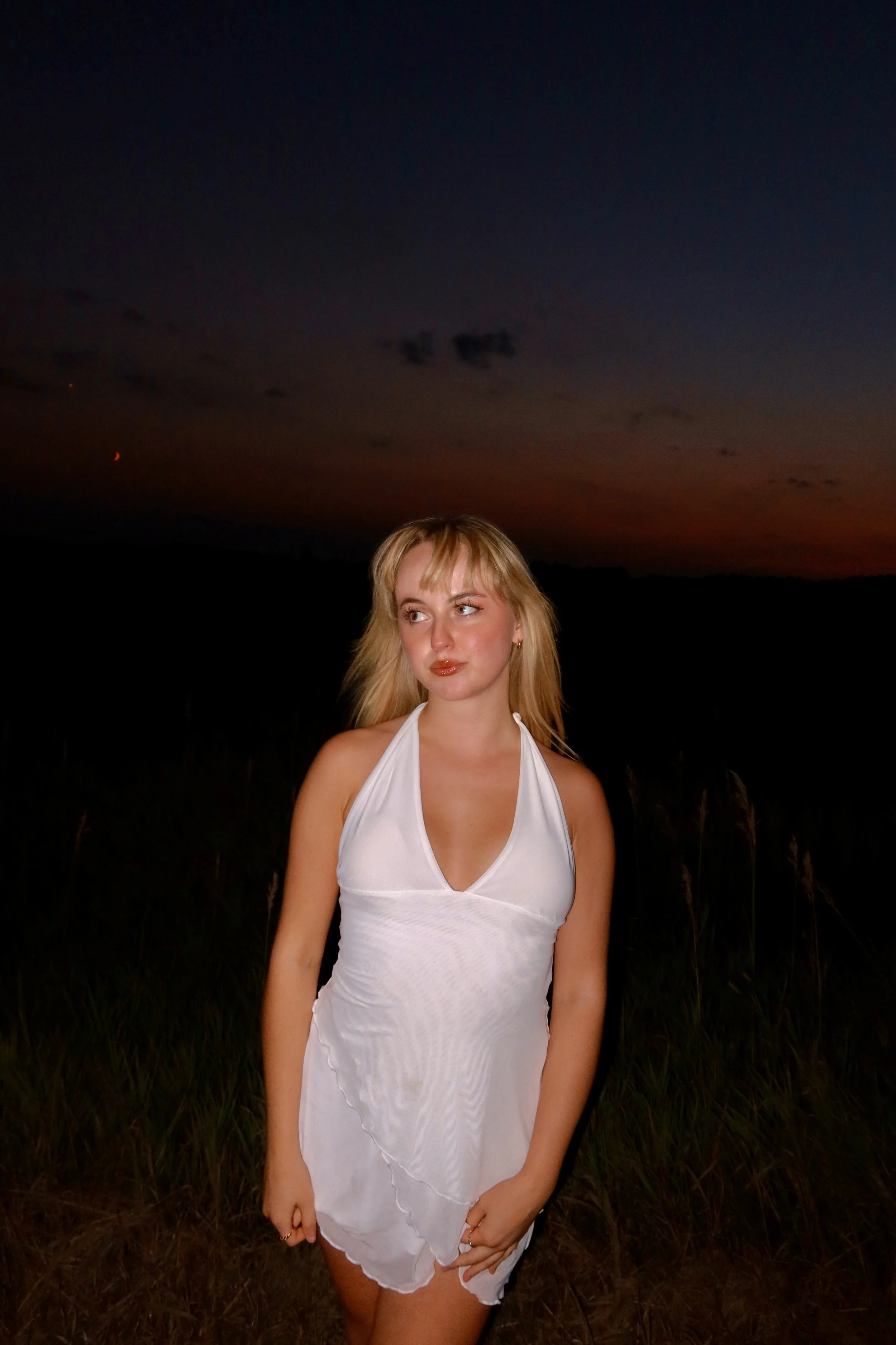 A young woman with blonde hair wearing a white dress standing outdoors at dusk or night. The sky behind her is dark with faint hints of sunset colors near the horizon.