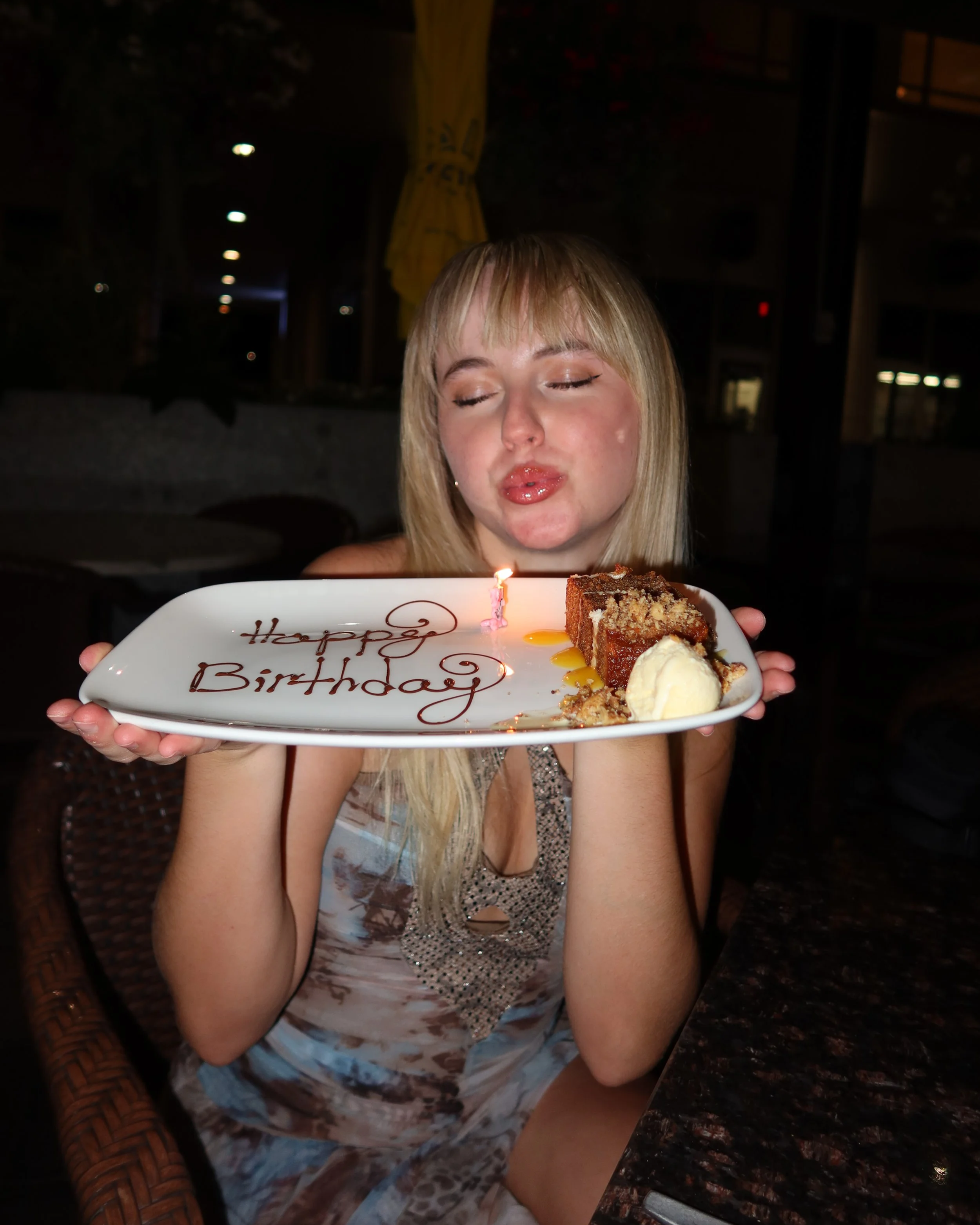 A woman with blonde hair and makeup holds a birthday plate with cake, ice cream, and a lit birthday candle, celebrating birthday.