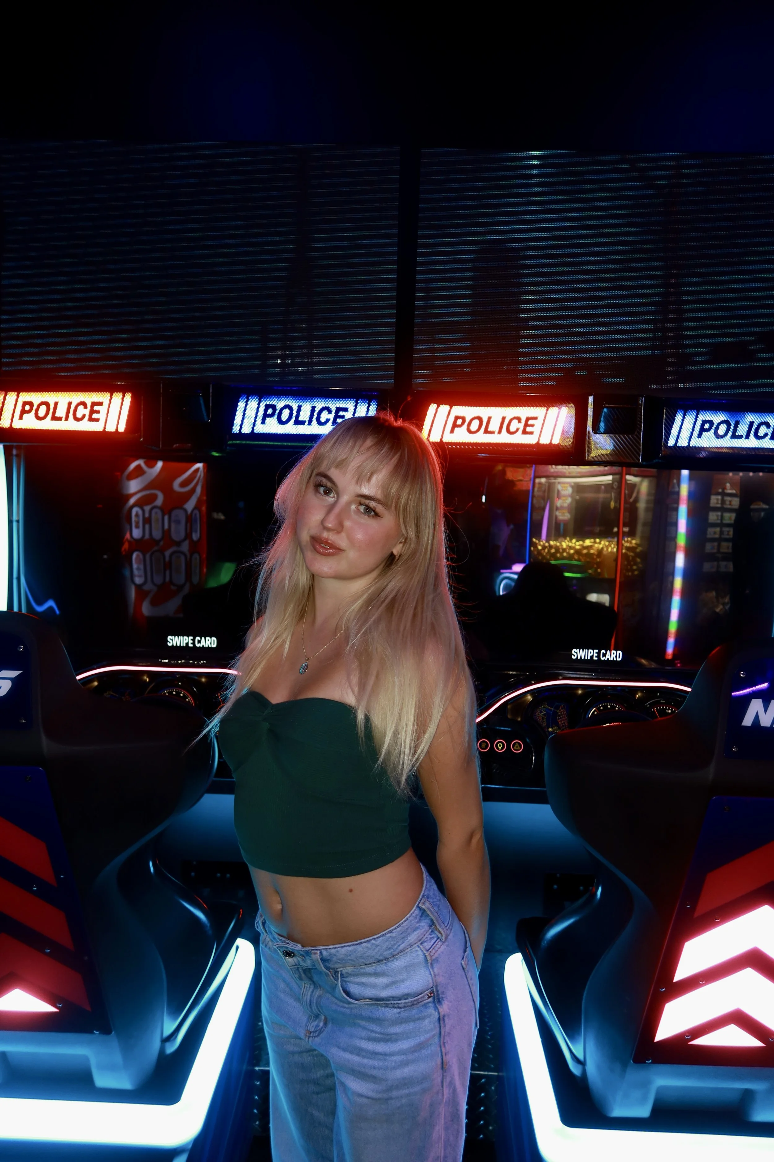 A young woman with blonde hair wearing a dark green crop top and light blue jeans standing inside an arcade with glowing neon lights, arcade game machines, and police-themed racing game cars.