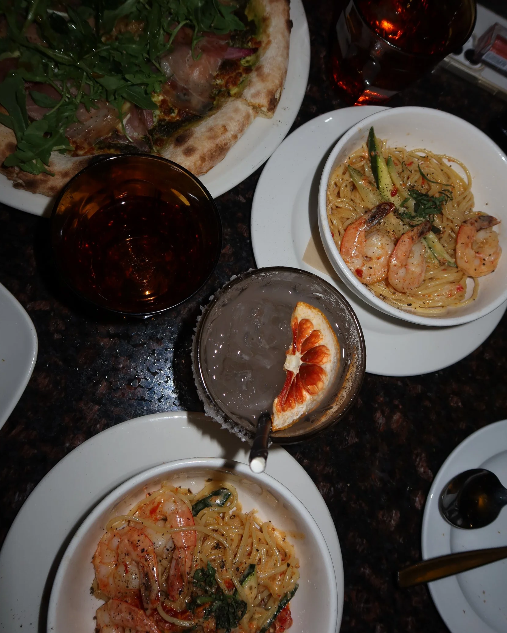 A table with a pizza topped with arugula, plus two glasses of dark soda, and two bowls of pasta with shrimp and sliced vegetables.