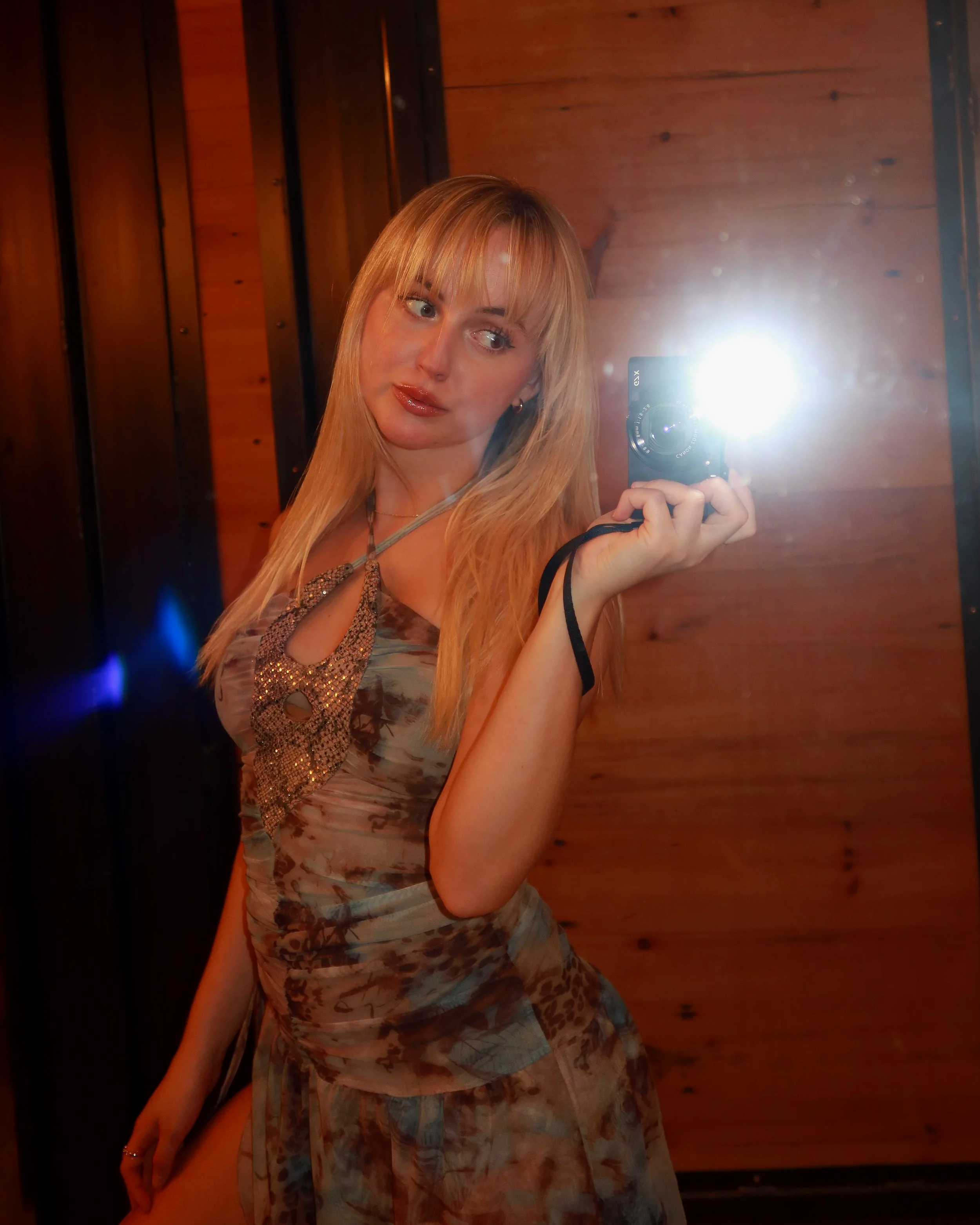 A woman with long blonde hair, wearing a floral dress and a necklace, taking a selfie in front of a wooden wall with warm lighting and a mirror.
