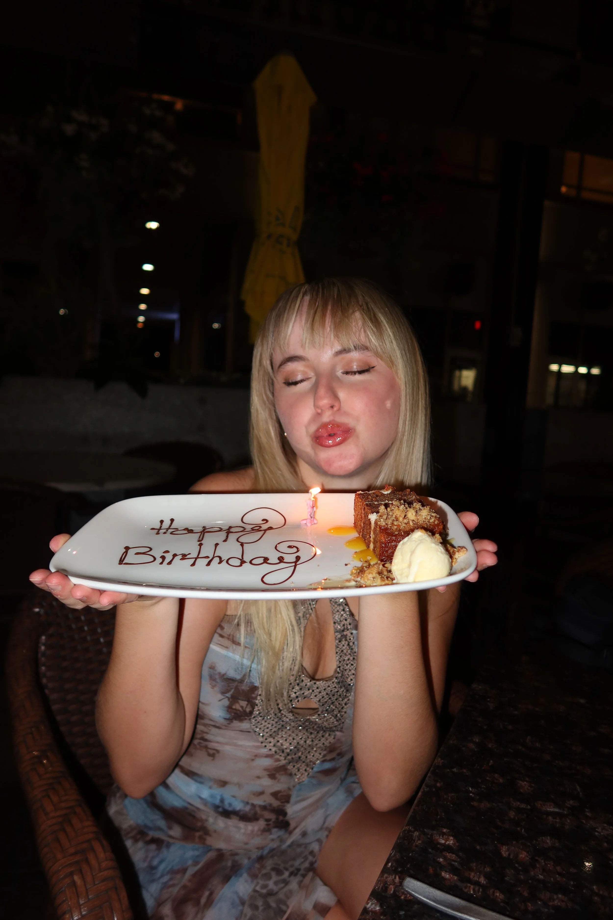 A young woman with blonde hair holding a birthday dessert plate with a slice of cake, a birthday candle, and ice cream. The plate says 'Happy Birthday' in chocolate script.