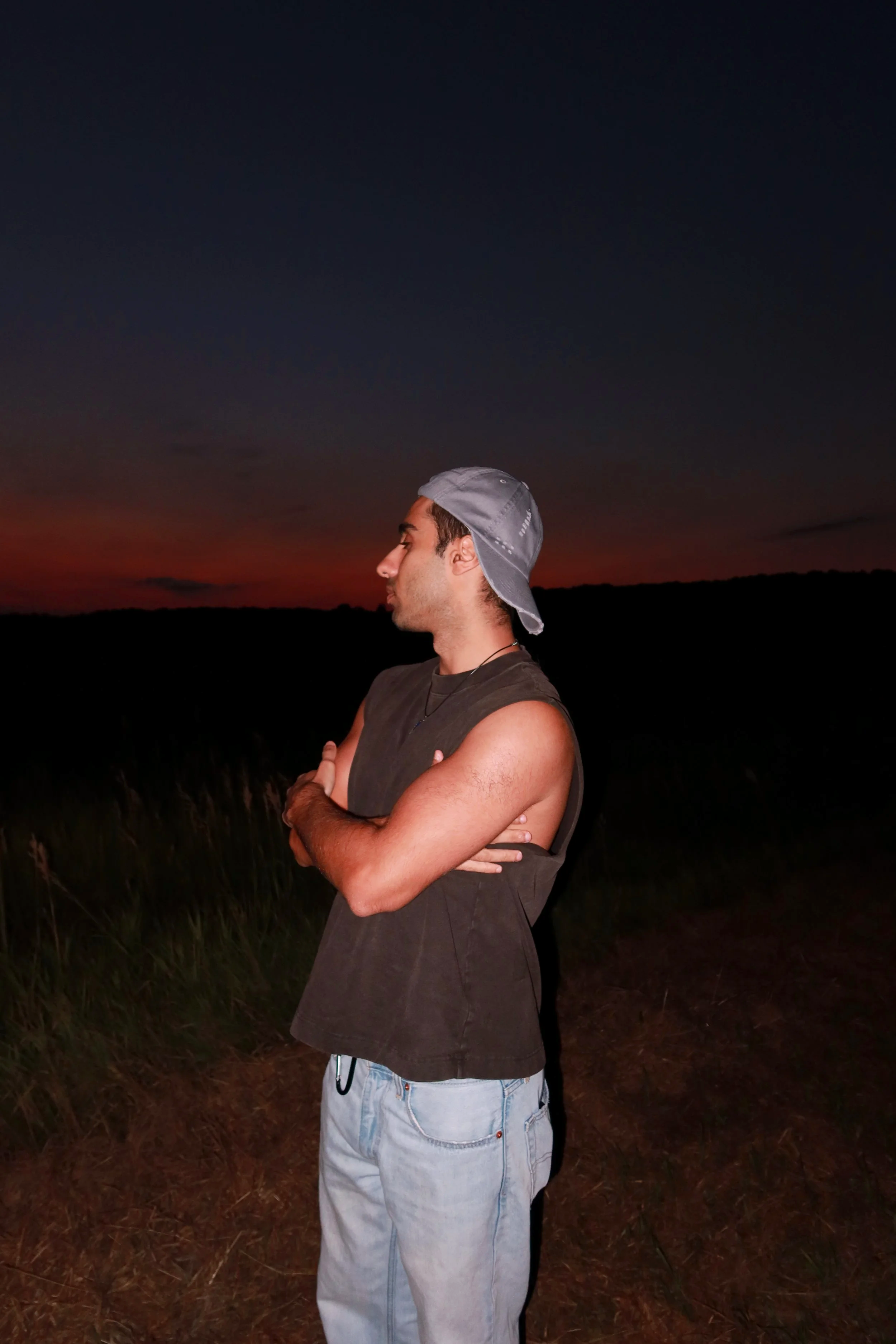 A young man with short dark hair wearing a gray baseball cap backwards, sleeveless dark t-shirt, and light-colored jeans standing outdoors during sunset with his eyes closed and arms crossed, in a natural setting.