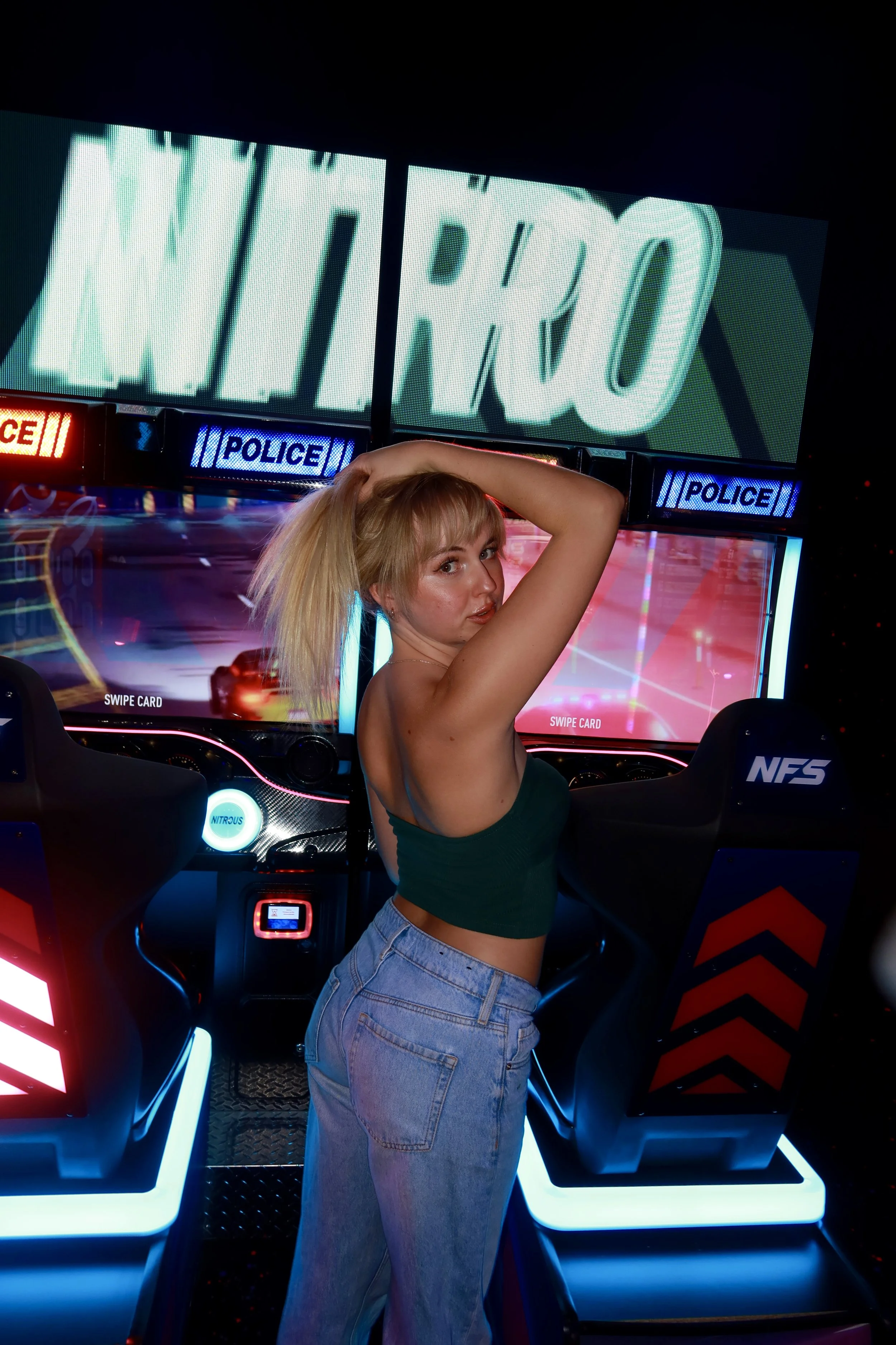 A woman with blonde hair, wearing a green tank top and blue jeans, leaning on an arcade racing game machine with neon lights, arcade controls, and a large screen displaying the word 'NIRRO' in the background.