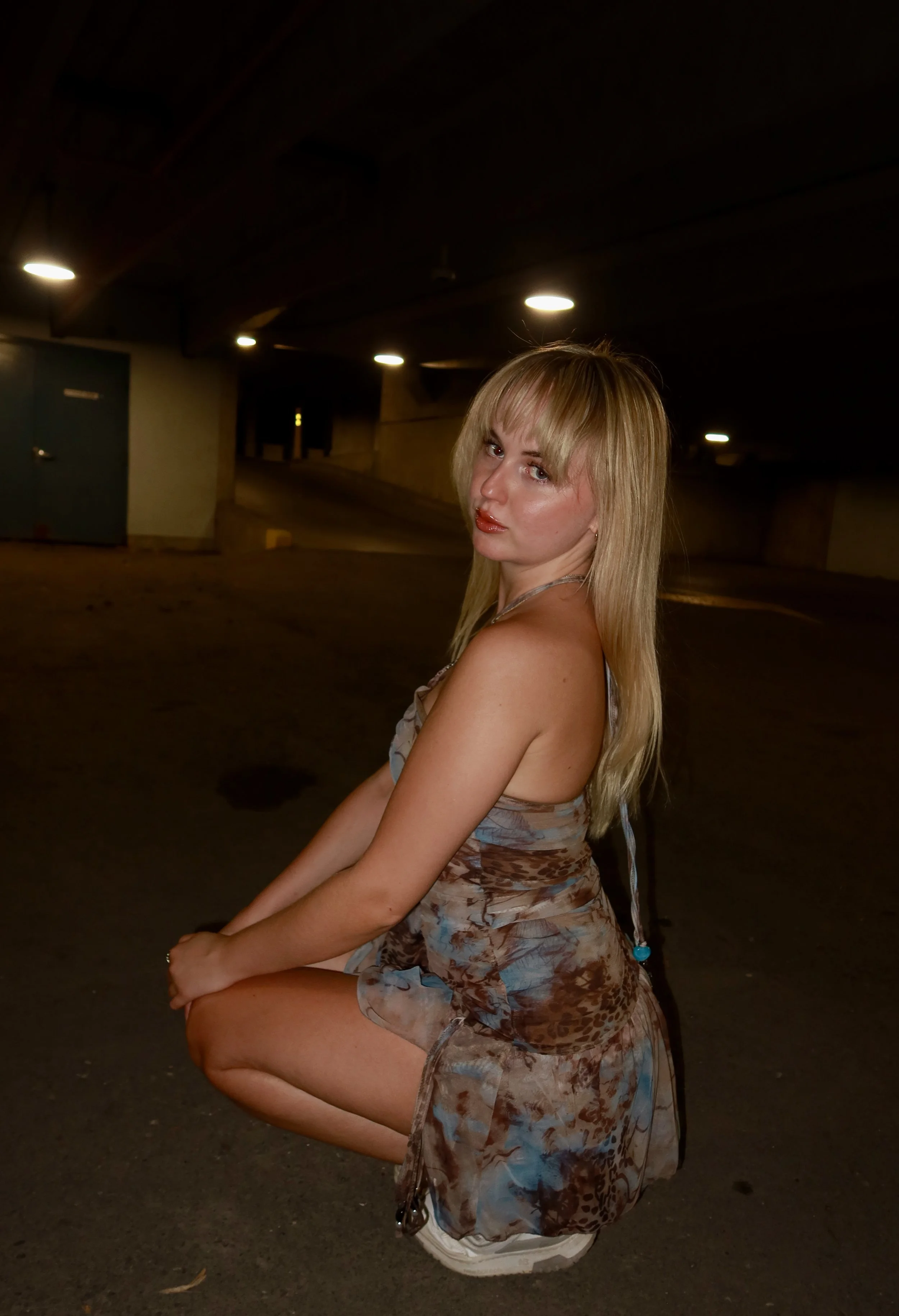 A young woman with long blonde hair and a patterned dress is crouching in a parking garage at night, looking over her shoulder at the camera.