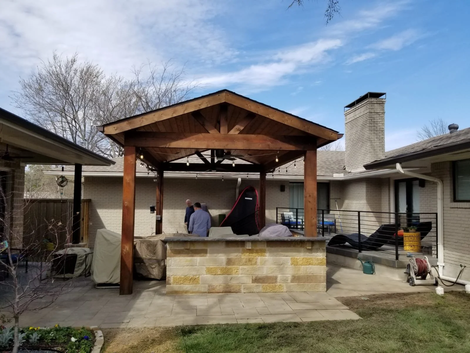 Backyard patio with a covered gazebo, two people talking, furniture including lounge chairs, a covered grill, countertop with a stone base, and gardening tools, enclosed by a fence and house wall, under the sunny yet cloudy sky.