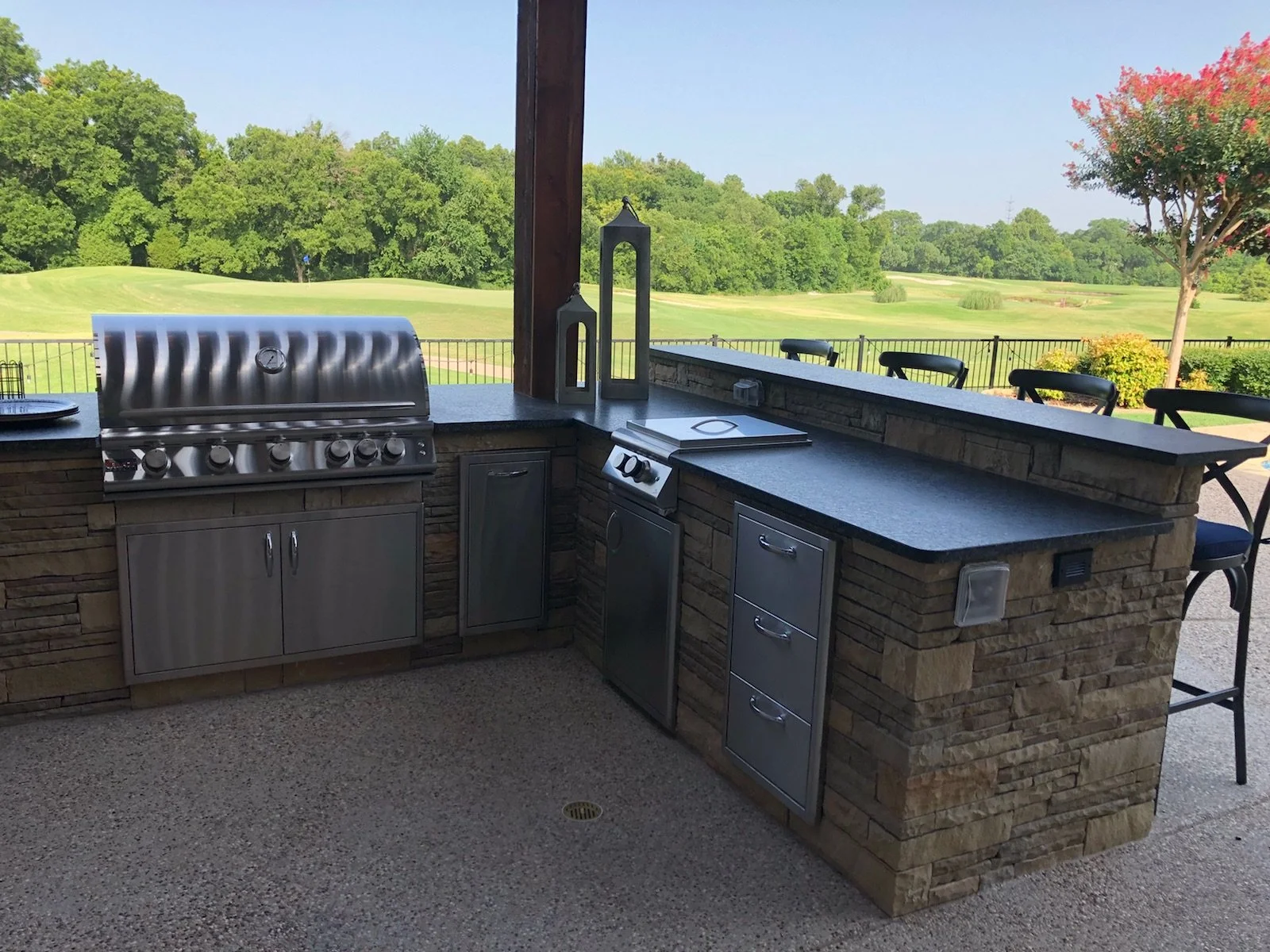 Covered outdoor kitchen with a built-in stainless steel grill, cabinets, and black countertop with four tall chairs behind it, overlooking a golf course with green trees and a blue sky.