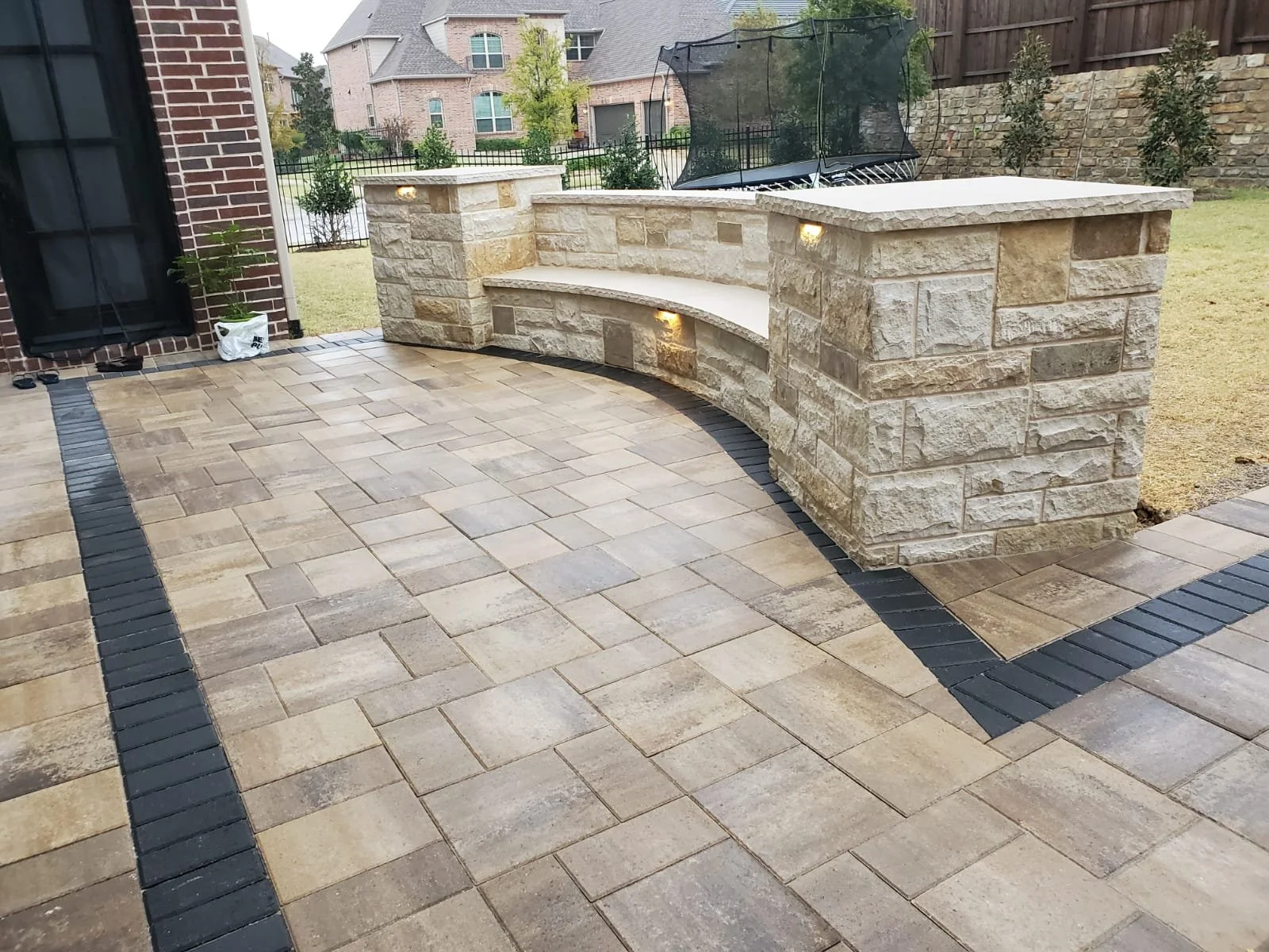 A backyard patio with tile work, a brick seating area with aesthetic lighting for nighttime, a trampoline, potted plants, and neighboring houses in the background.