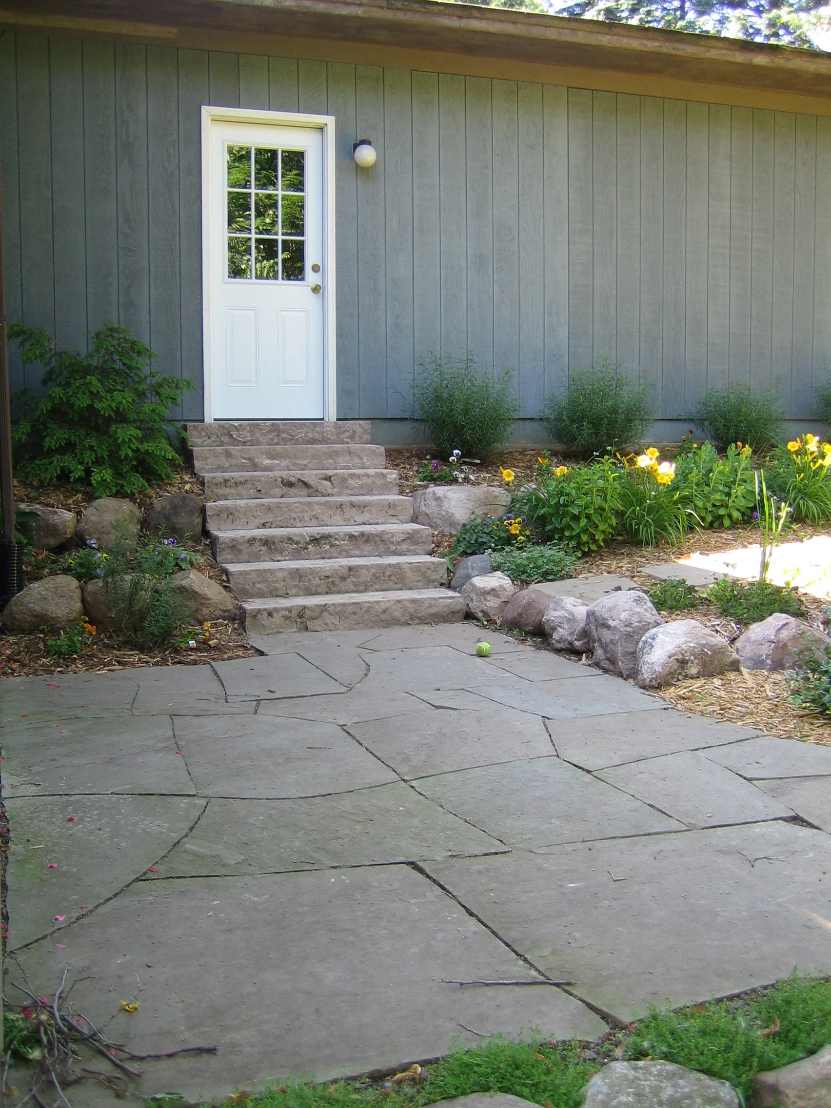A backyard patio with stonework and stone steps leading up to a white door on a gray wooden house, surrounded by greenery and beautifully arranged and planted flowers.