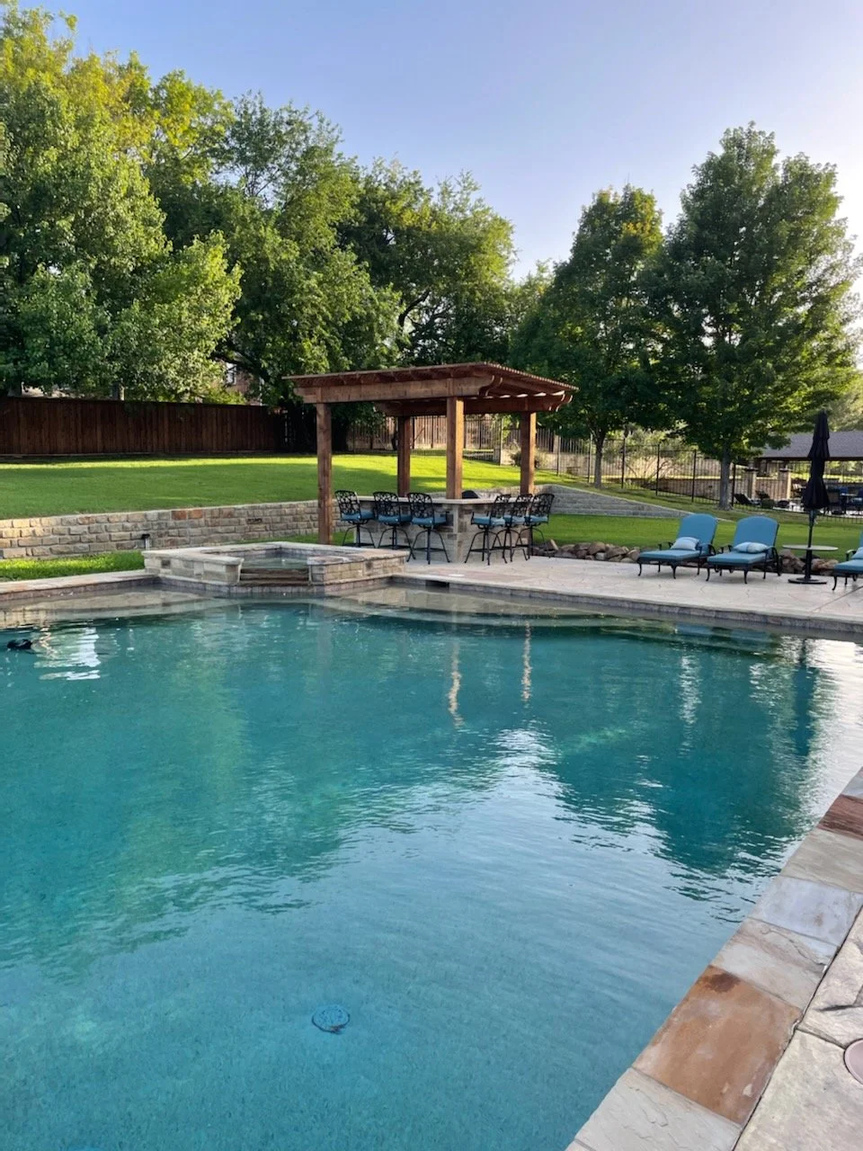 Residential swimming pool in backyard with shaded seating area, lounge chairs, and tall trees in the background.