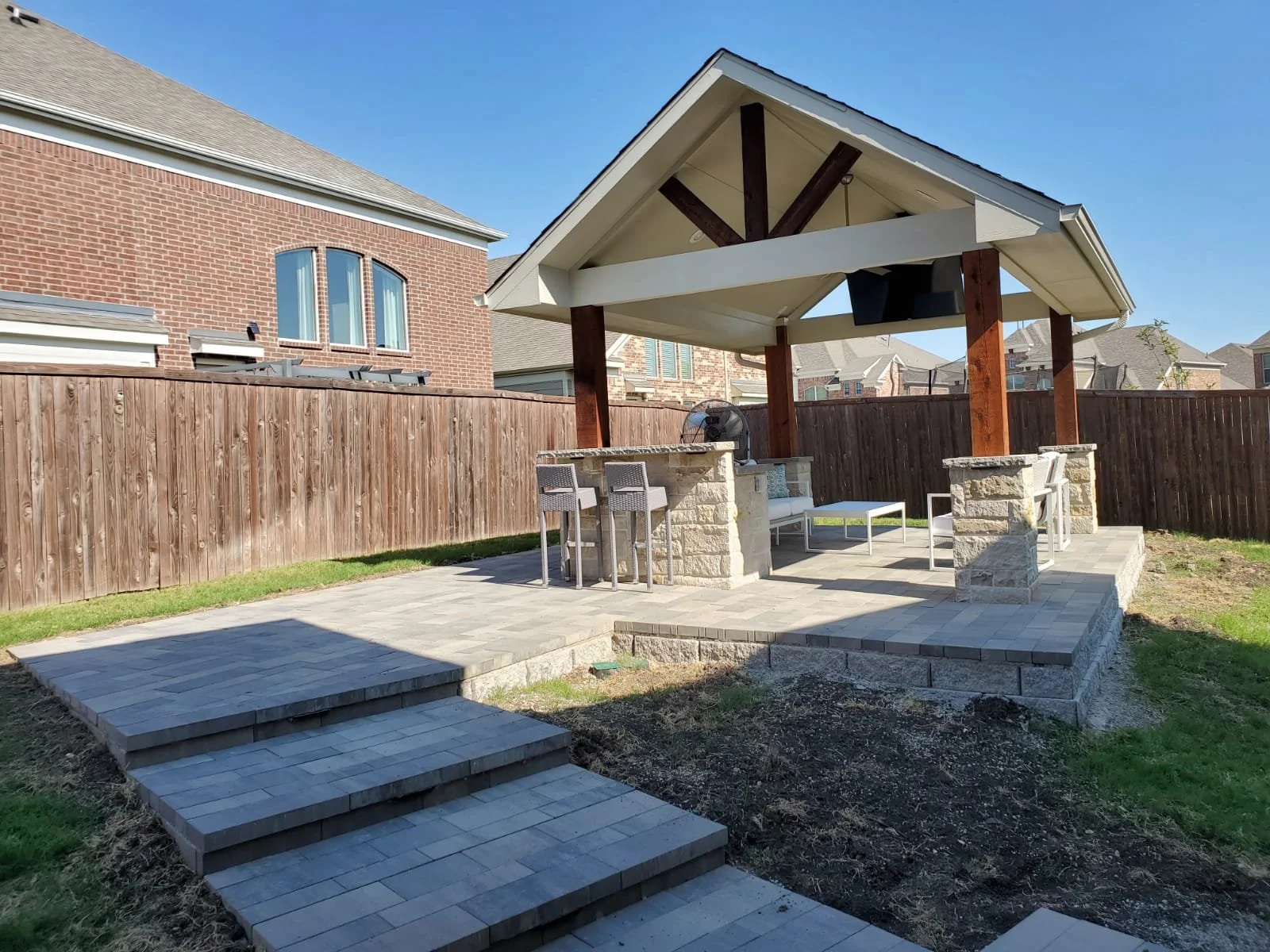 Backyard patio with a covered outdoor bar and seating area, brick and stone construction, wooden fence, in a suburban neighborhood.