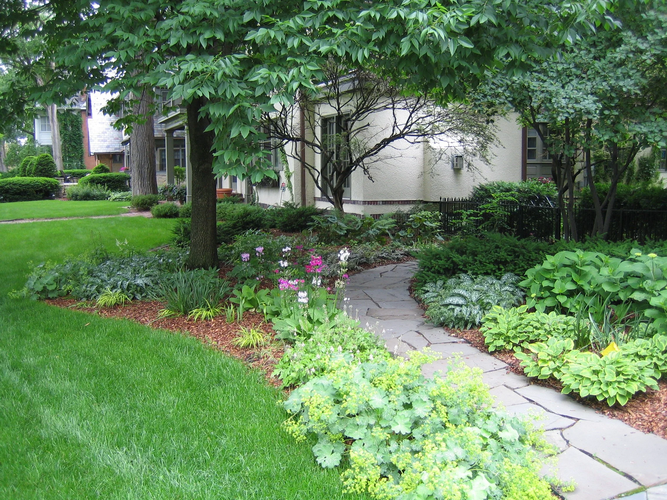A lush green garden with neatly trimmed grass, flowering plants, shrubs, and trees, leading to a house with a white exterior and a pathway.